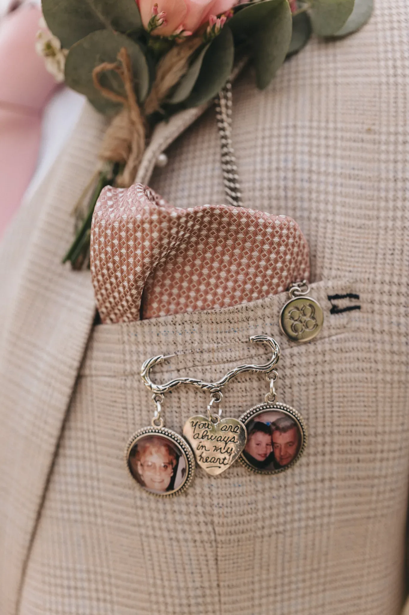 A close-up of a lapel pin on a tweed jacket featuring two photo charms and an engraved pendant that reads "you are always in my heart," beside a rose boutonniere. the photos in the charms depict smiling faces.