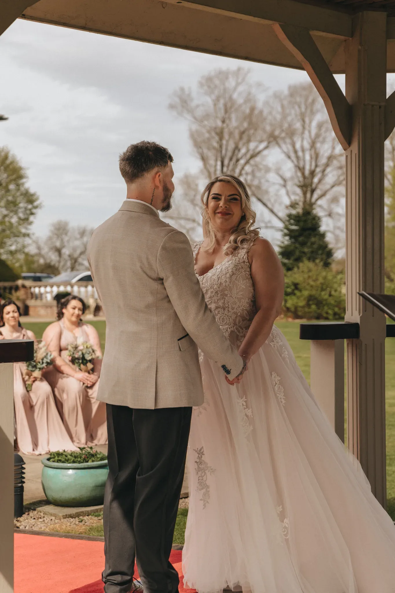 A bride and groom stand under a wooden gazebo, holding hands and smiling at each other. the bride is wearing a detailed white wedding dress, and the groom is in a light grey suit. bridesmaids in pink dresses are visible in the background.