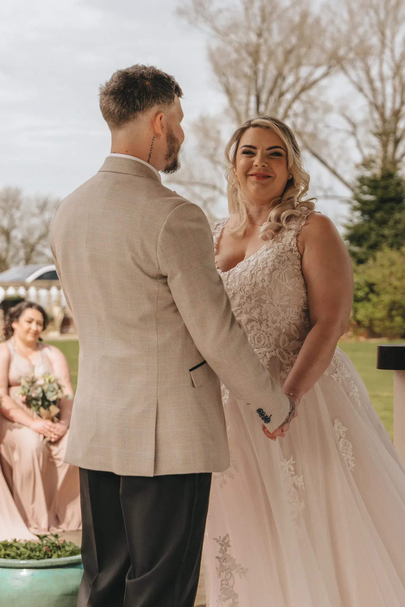 A bride and groom stand facing each other during a wedding ceremony. the bride, in a lace gown, smiles while the groom, in a beige suit, stands with his back to the camera. a bridesmaid in a pink dress observes from behind.