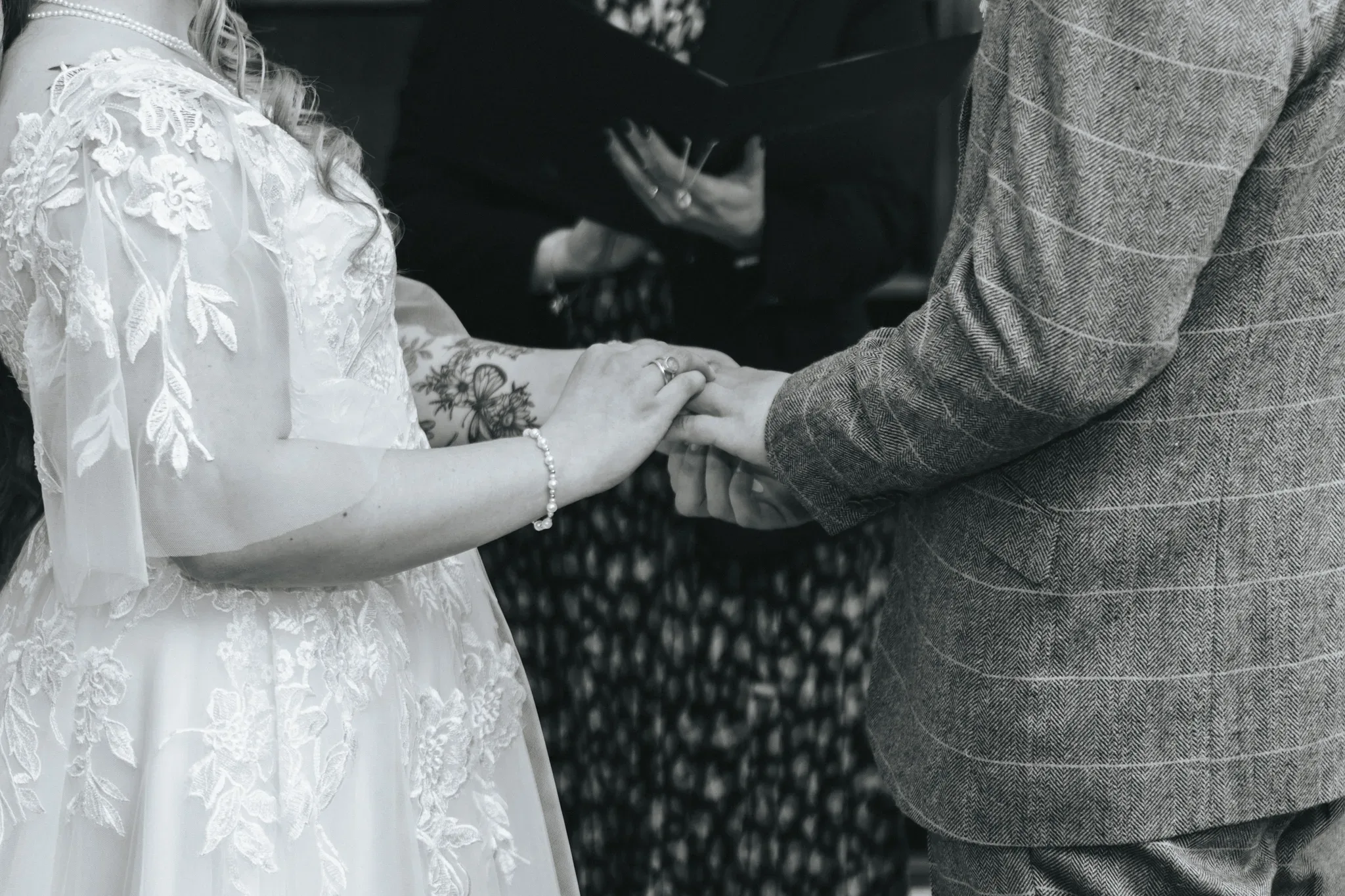A bride and groom hold hands during a wedding ceremony. the bride, in a white dress with lace sleeves, displays a tattoo on her arm, while the groom wears a gray tweed suit. a person officiating the ceremony stands in the background.