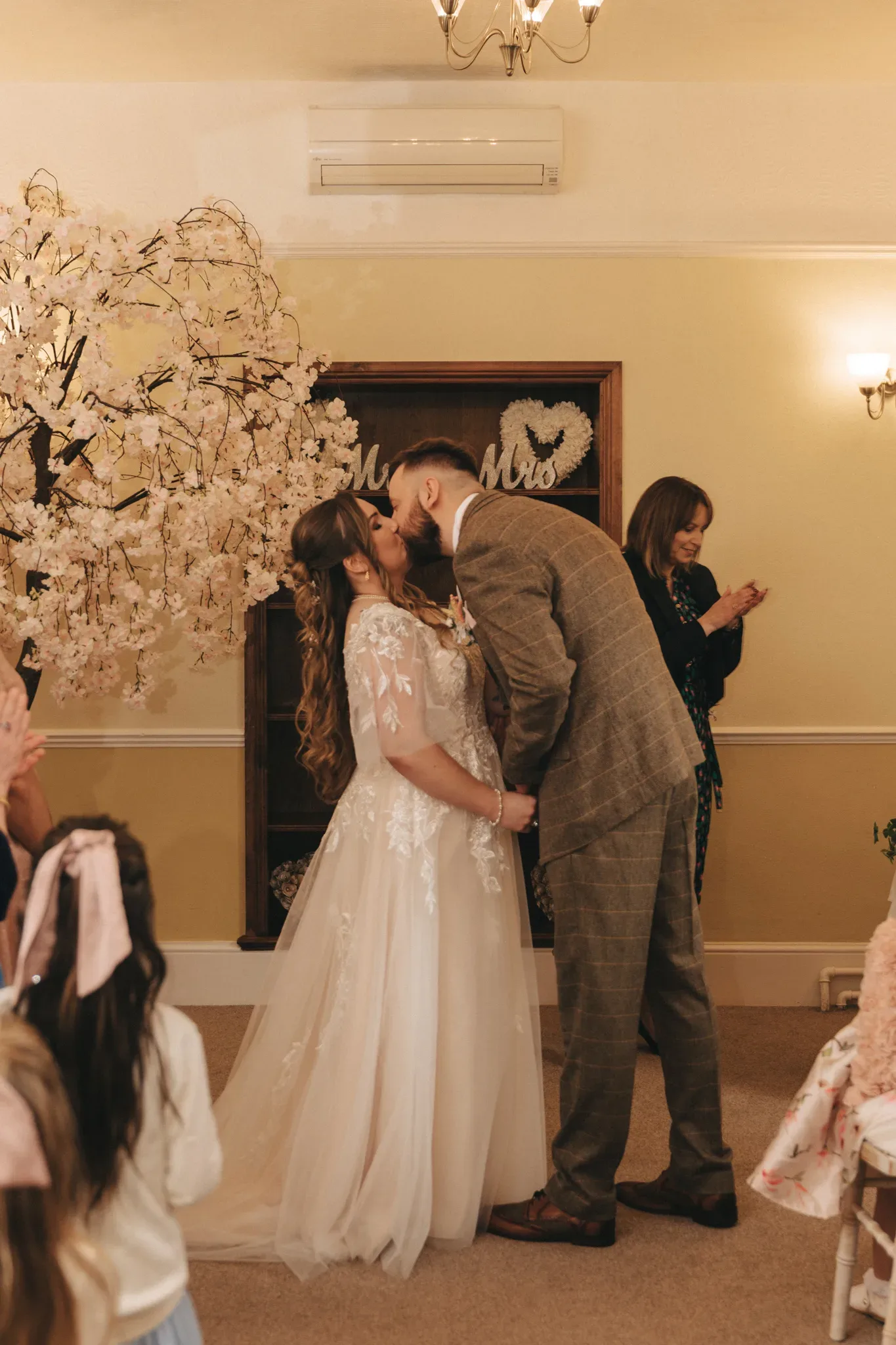 A bride and groom share a kiss under a cherry blossom tree indoors, surrounded by guests. the bride is in a lace gown, and the groom wears a tweed suit. decor includes "mr & mrs" signs.