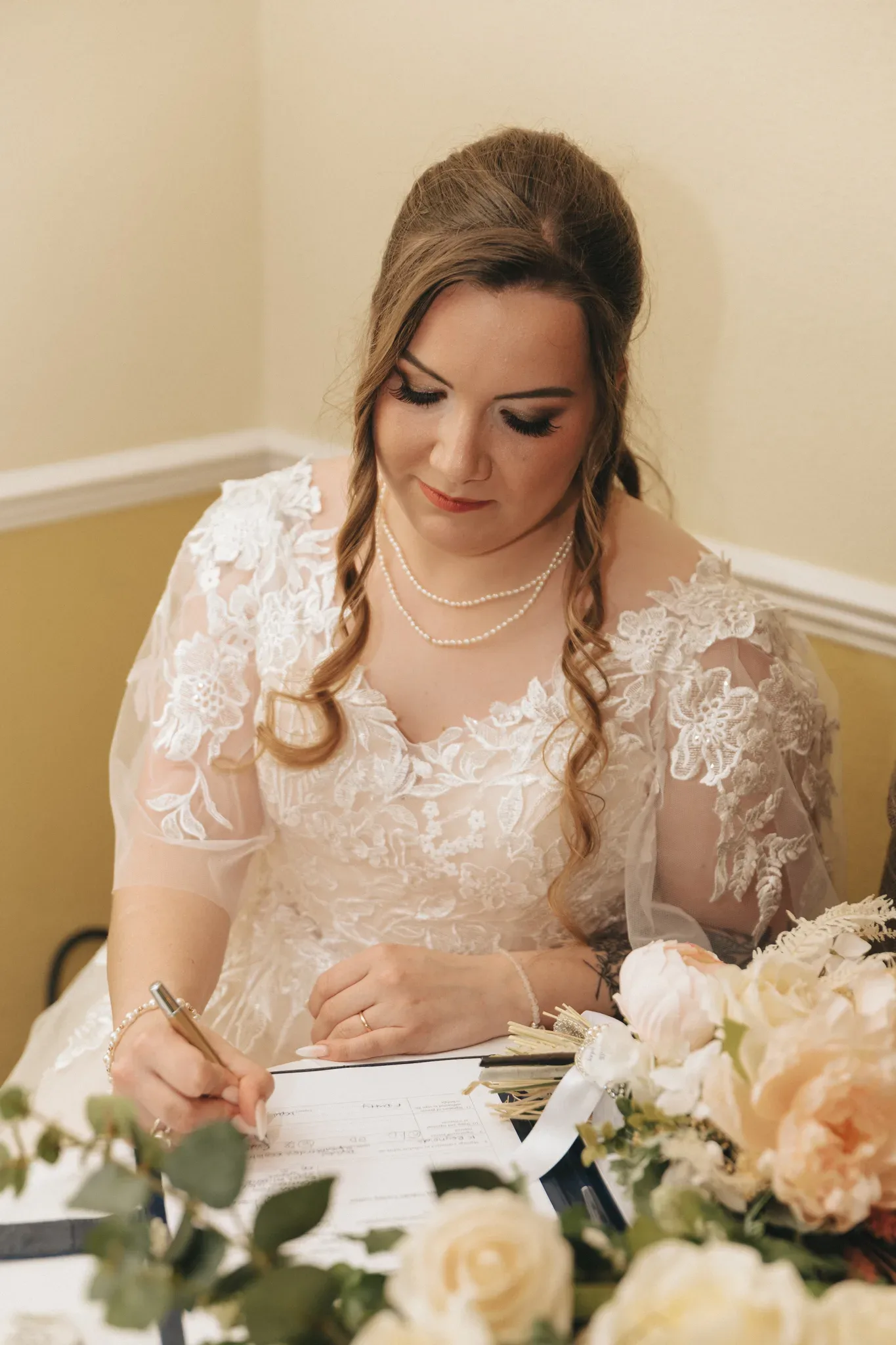 A bride in a lace dress and pearl necklace signs a document at a table adorned with floral bouquets, looking down with a gentle smile in a warmly lit room.