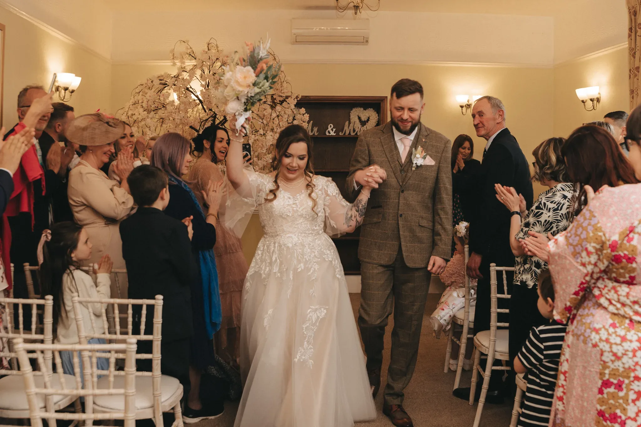 A newlywed couple joyfully exits their wedding ceremony, walking hand-in-hand through a crowd of applauding guests. the bride, in an elegant white dress, and the groom, in a tweed suit, smile broadly.