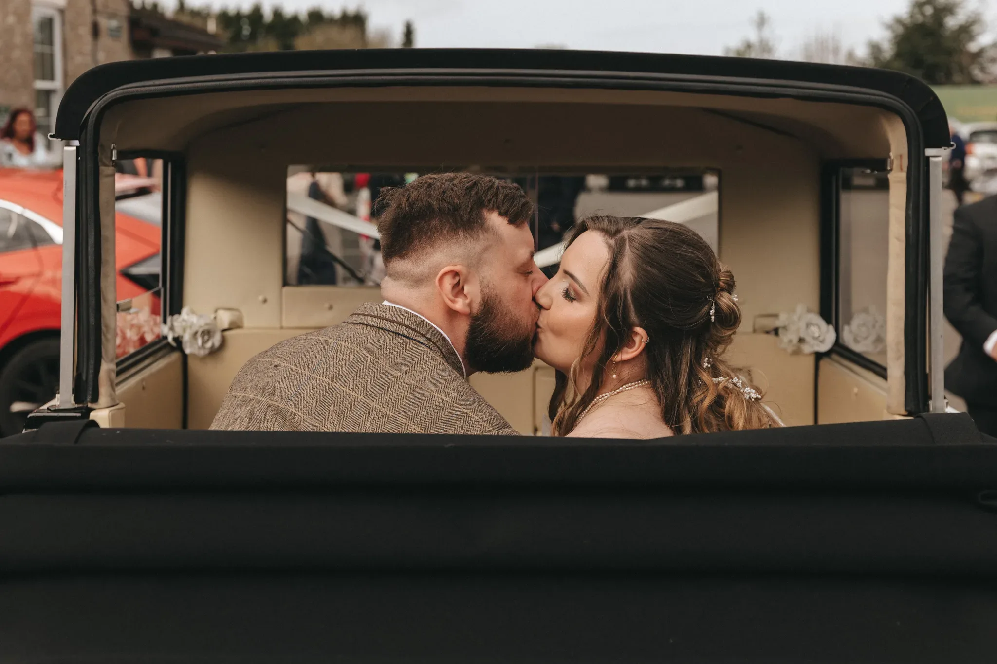 A bride and groom share a kiss inside a vintage car with the top down. the background shows a softly blurred hillside and other vintage cars, creating a romantic, timeless atmosphere.