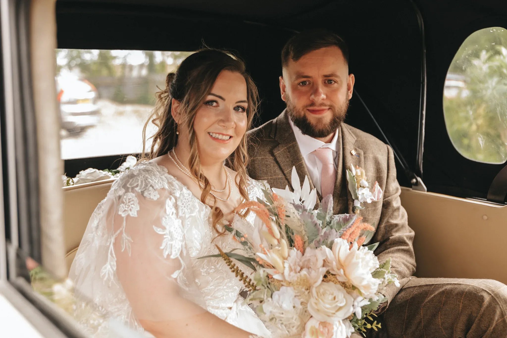 A bride and groom smiling inside a vintage car, the bride in a lacy dress holding a bouquet of white and peach flowers, the groom in a tweed suit with a pink tie.