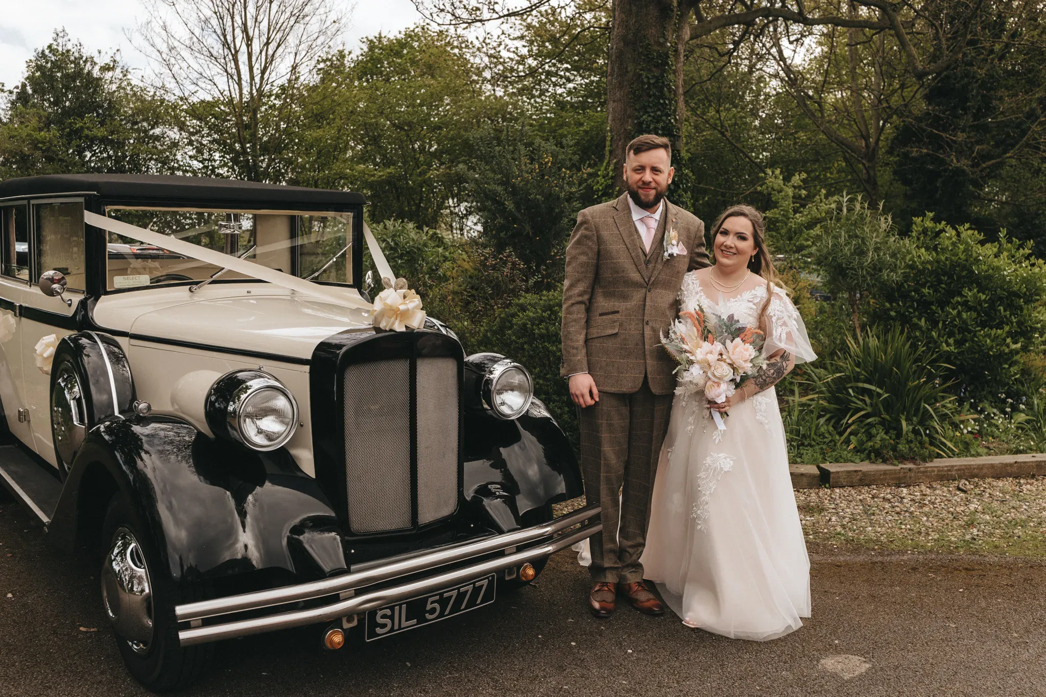 A bride and groom stand beside a vintage car in a park. the groom wears a tailored brown suit; the bride, a white gown with a bouquet. the car is black and white, adorned with a flower arrangement.