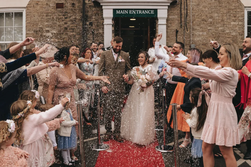 A joyful bride and groom exit a building through a crowd of friends and family who are throwing confetti. the scene is festive, with guests in colorful attire capturing the moment, and a red carpet under the couple's feet.