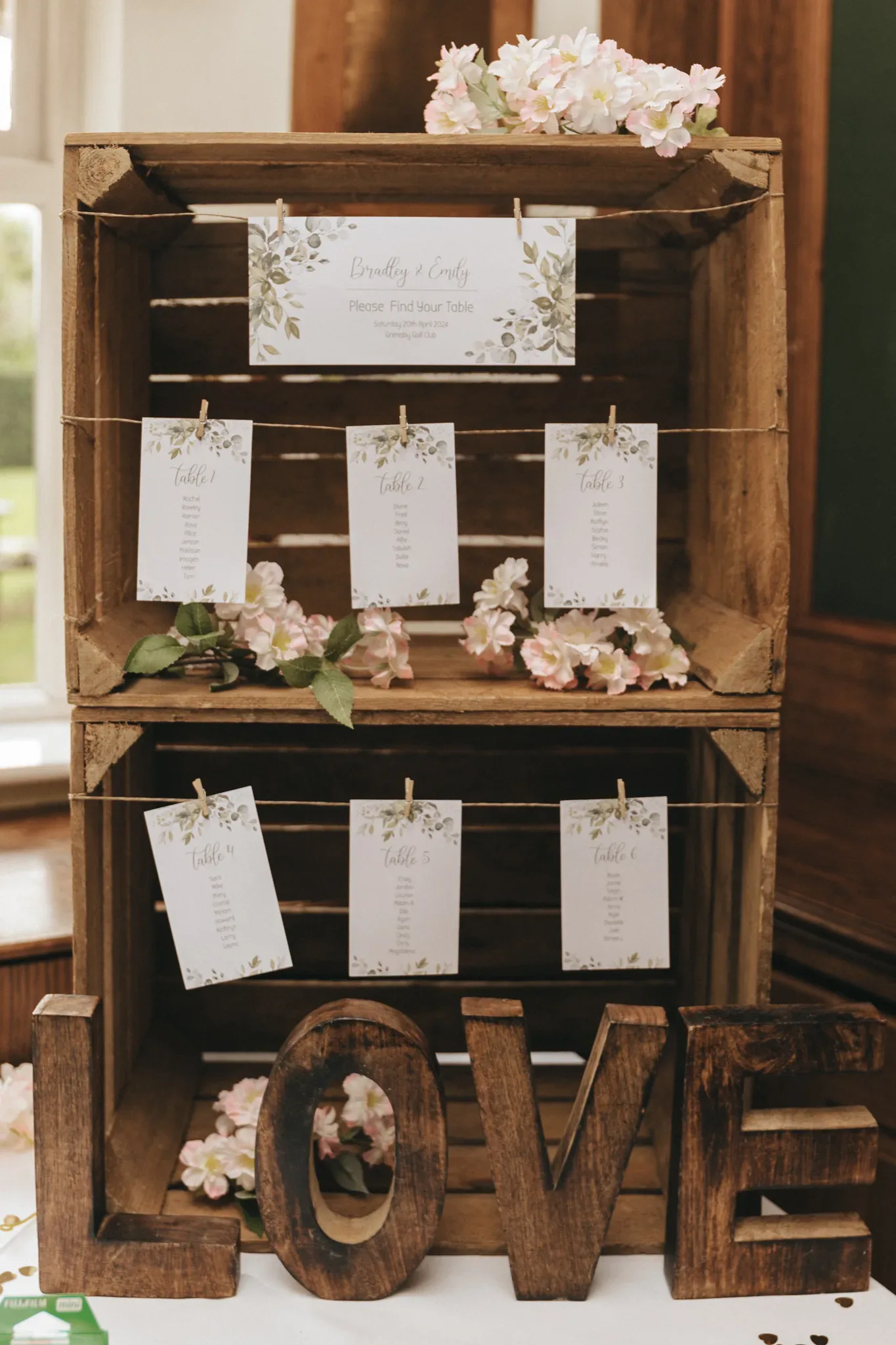 A rustic wooden crate displays wedding seating arrangement cards, adorned with floral decorations. a 'love' decoration in large letters is prominently featured at the bottom, alongside a floral-framed mirror with a reflection of flowers.