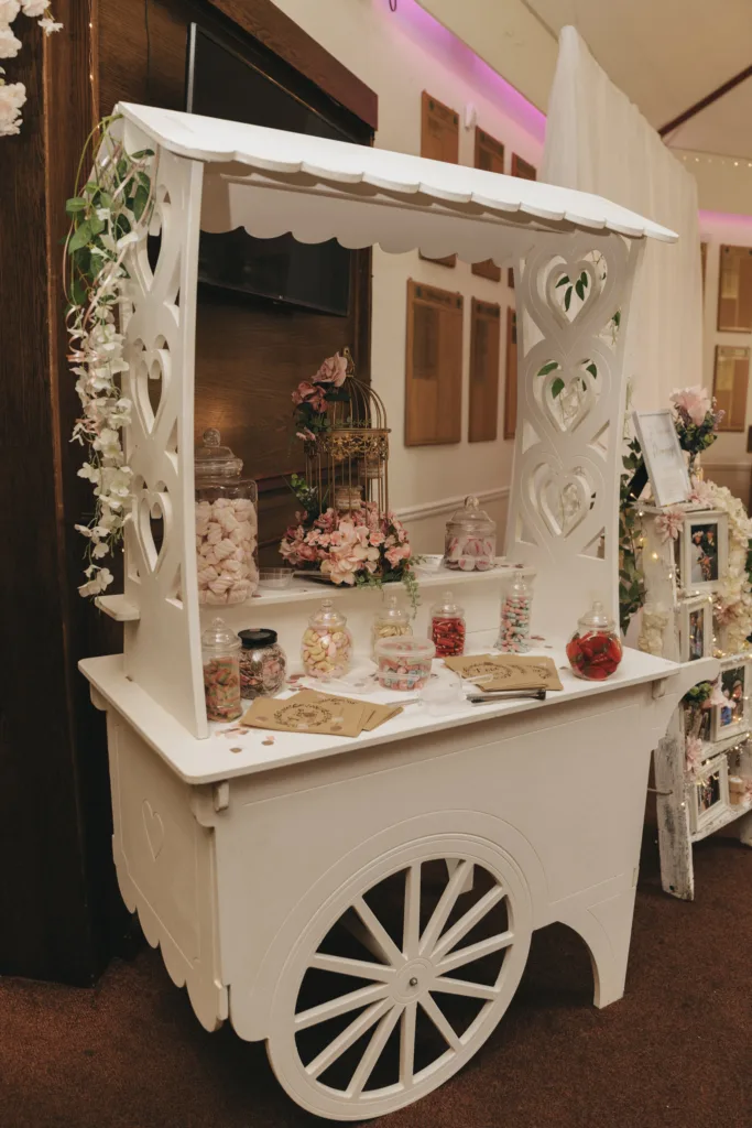 A charming white candy cart decorated with flowers, featuring various jars of colorful candies and cookies, set up indoors with warm lighting for a festive or wedding event.