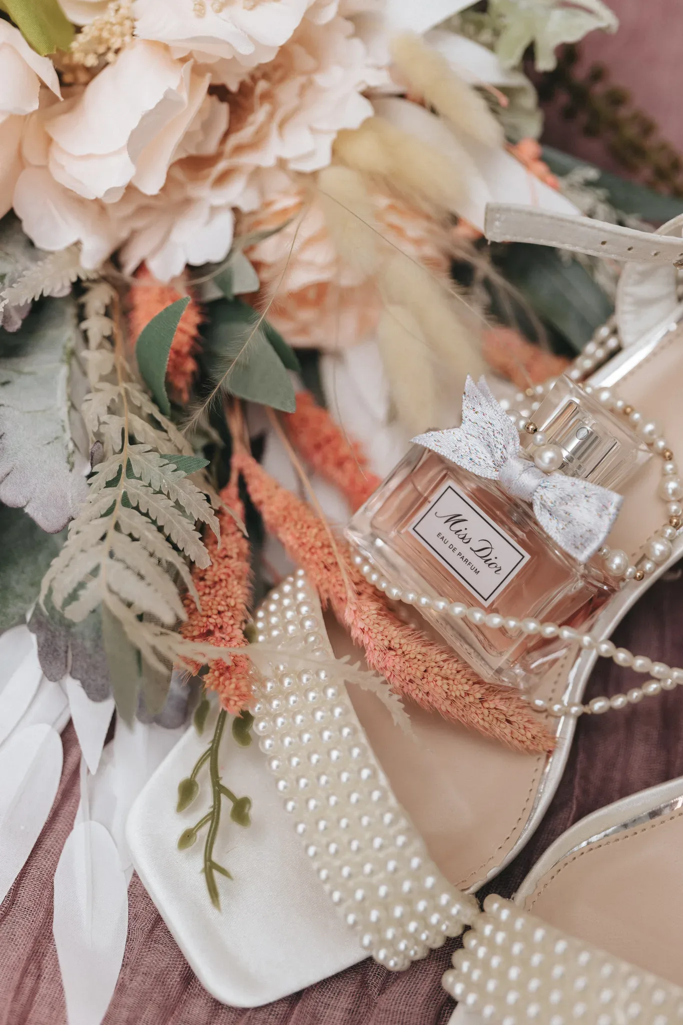 A close-up image of a perfume bottle labeled "miss dior" placed on women's shoes adorned with pearls and surrounded by vibrant floral arrangements and greenery on a textured pink surface.