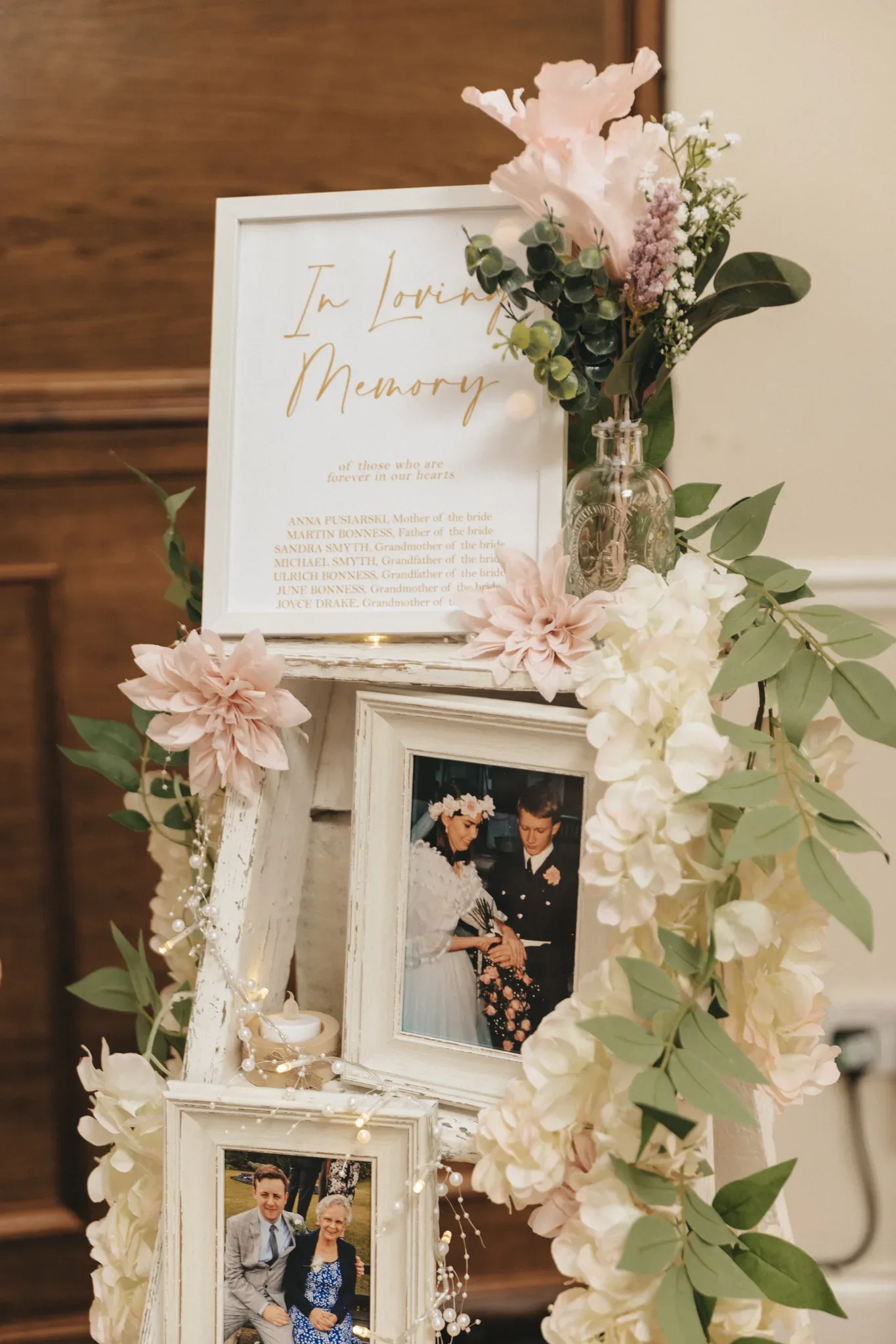 A memorial display features a floral arrangement and photos within a white, tiered stand. a sign reading "in loving memory" lists names of the deceased. the decor includes soft pink flowers and string lights.