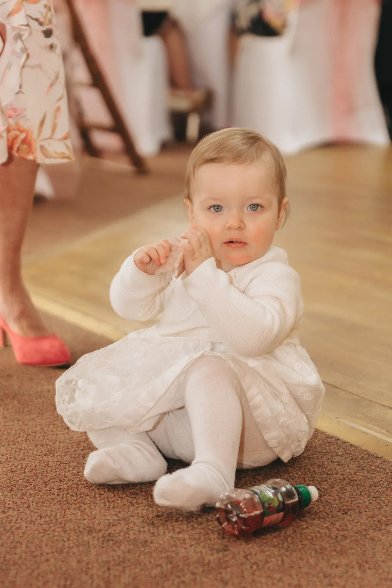 A toddler in a white dress and tights sits on a wooden floor, holding their hands together. the child has blue eyes and light brown hair, with a colorful toy nearby. a partial view of two adults is in the background.