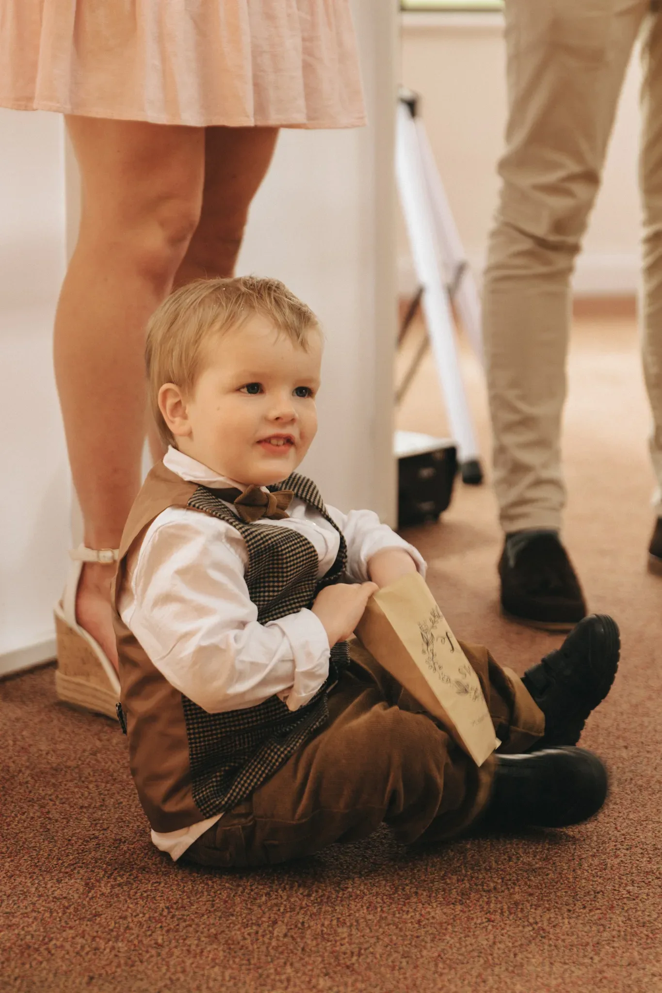 A toddler boy sits on the floor smiling, dressed in formal attire with a bow tie and suspenders. he is surrounded by standing adults, partially visible, focusing attention on him.