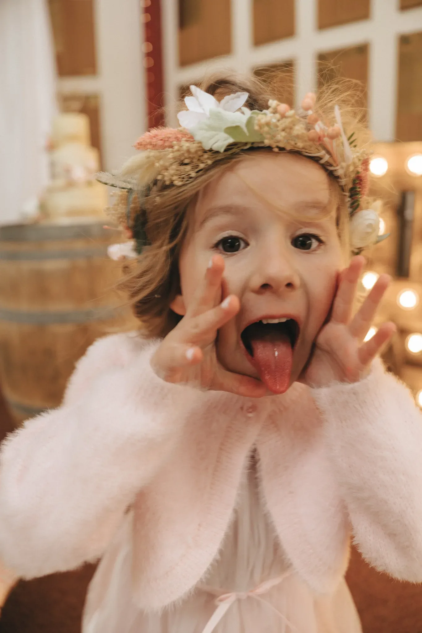 A young girl with a flower crown pulls a playful face by stretching her mouth with her fingers and sticking out her tongue. she wears a fluffy, light pink coat and stands indoors with warm lighting in the background.