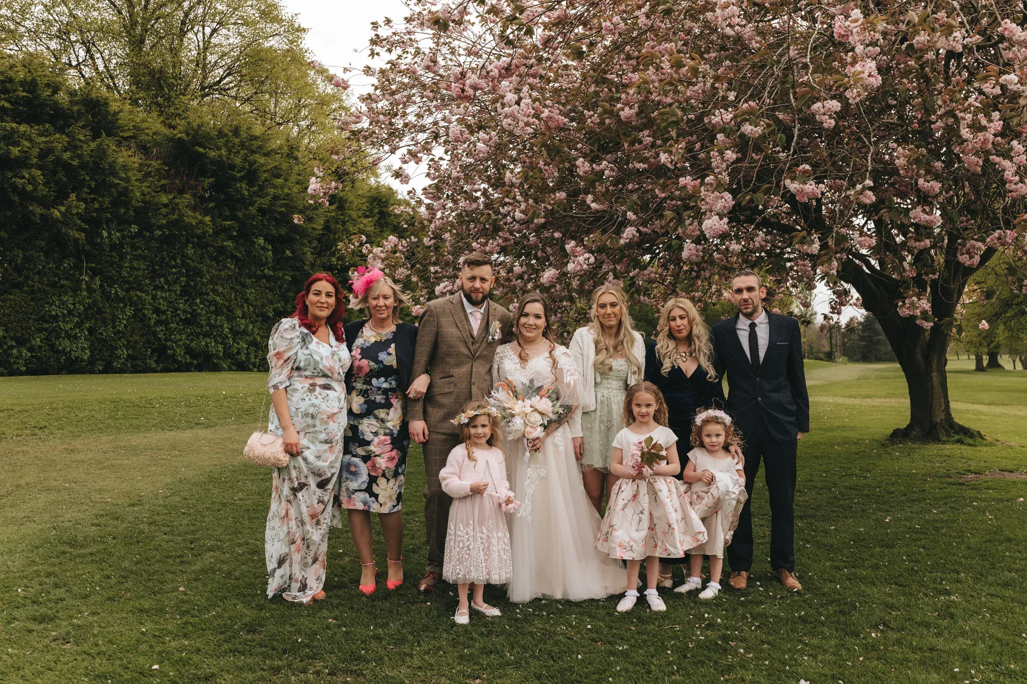 A wedding group photo under a blooming cherry blossom tree. the bride and groom are centered among nine family members and friends, wearing elegant dresses and suits, with the women holding bouquets. the setting is a lush green park.