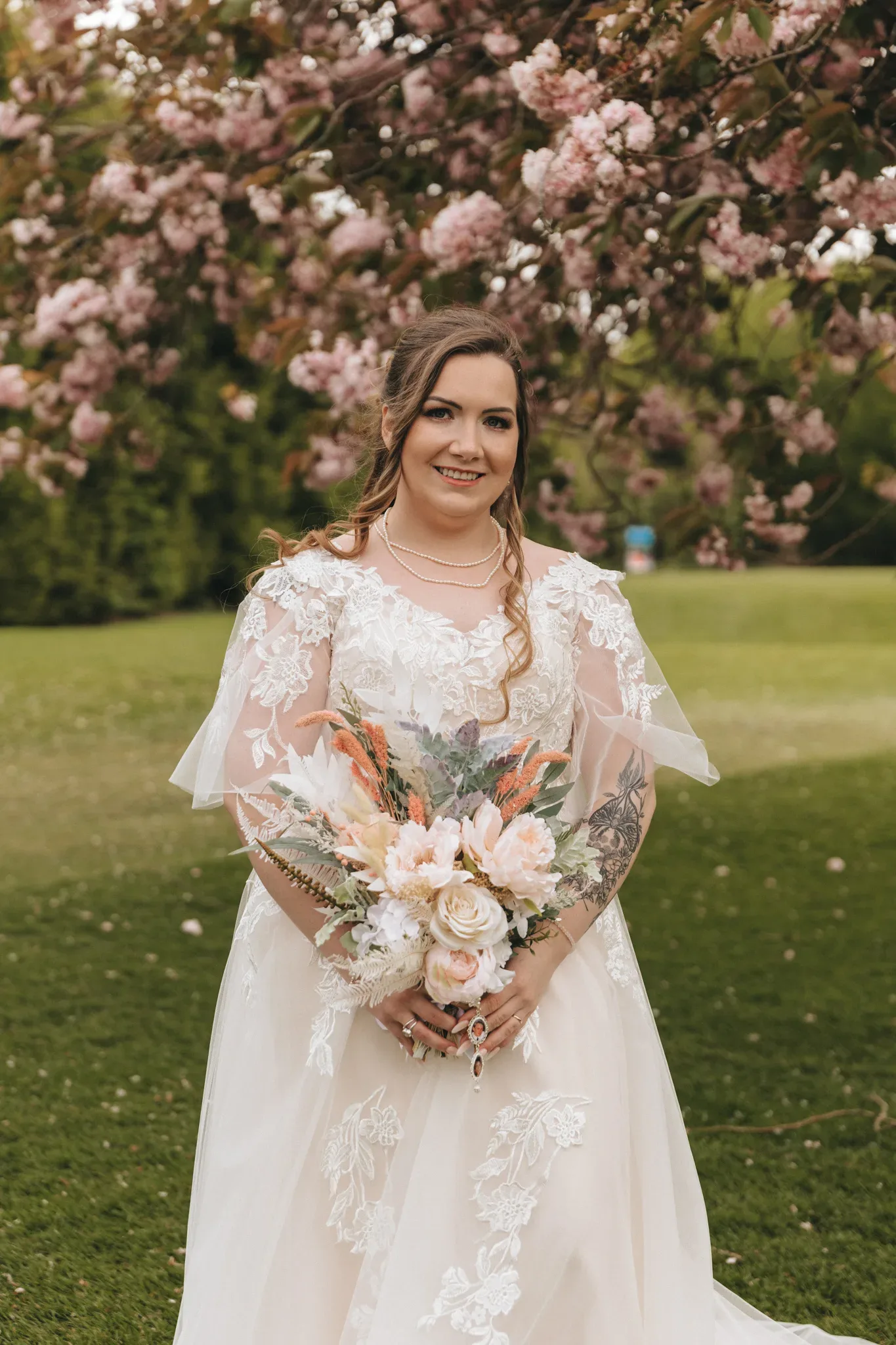 A bride stands under a blooming cherry tree, smiling at the camera. she wears a lace wedding dress and holds a bouquet of pink and peach flowers. her tattooed arms are partially visible.