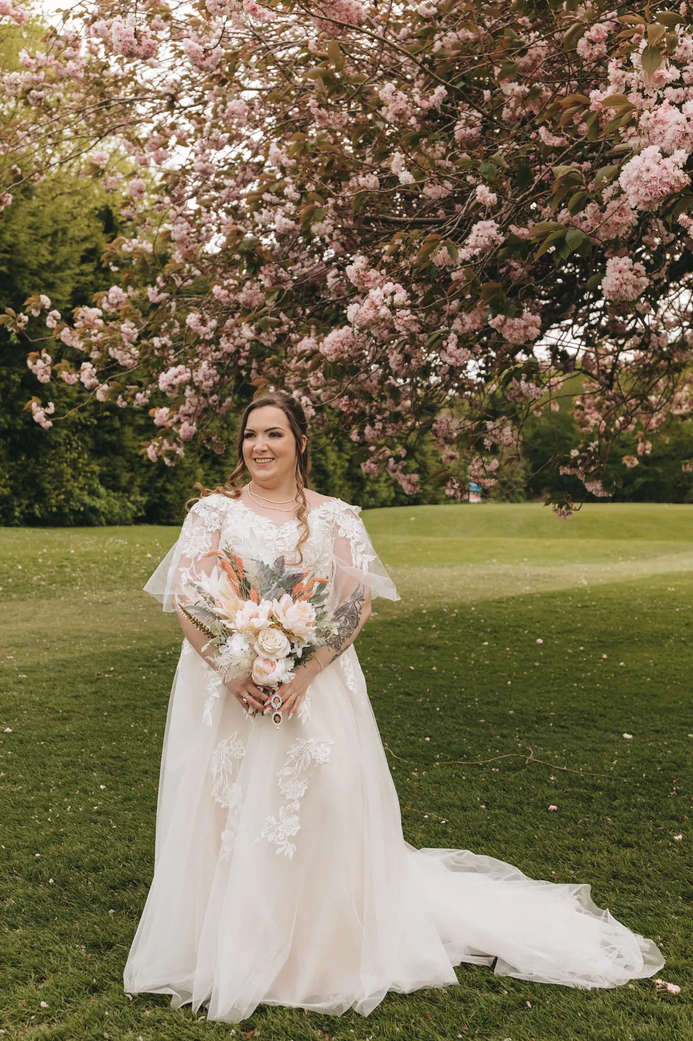 A bride in a lace wedding dress smiles under a blossoming pink tree, holding a large bouquet with pink and orange flowers. she stands on a grassy field, conveying joy and elegance amidst a serene natural setting.