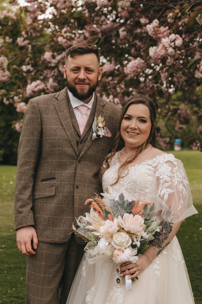 A bride and groom smiling and standing together under a blooming cherry blossom tree. the bride holds a bouquet of peach and cream flowers, and both wear traditional wedding attire.