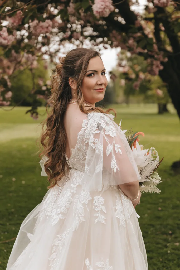 A bride with long, curled hair and wearing a detailed lace wedding dress holds a bouquet, looking over her shoulder under a canopy of pink blossoming trees in a lush park.