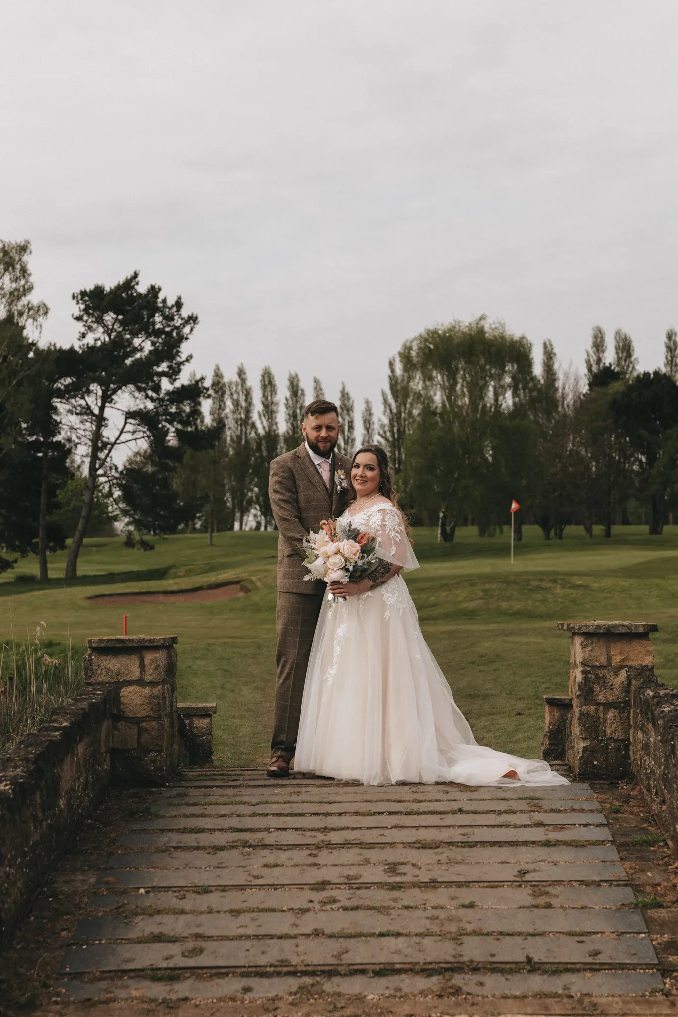 A bride in a white dress and a groom in a gray suit stand on a stone bridge in a park. the bride holds a bouquet and they both smile at the camera, with green trees and a cloudy sky in the background.