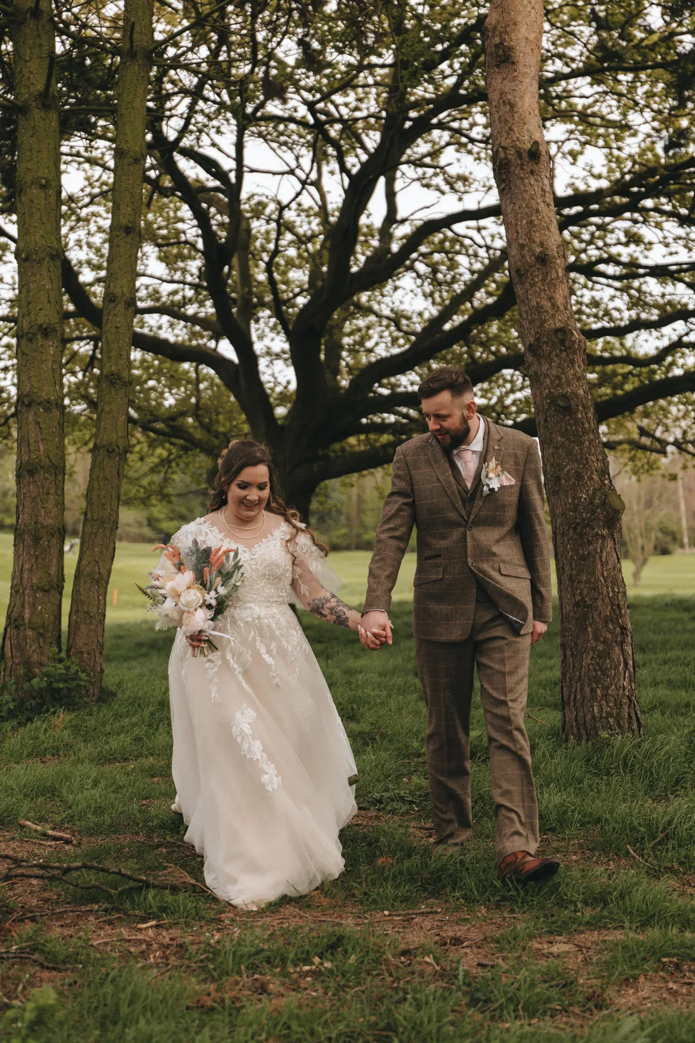 A bride and groom walk hand in hand in a grassy park with large trees, smiling joyfully. the bride wears a white lace gown and holds a bouquet, while the groom is in a tweed suit.