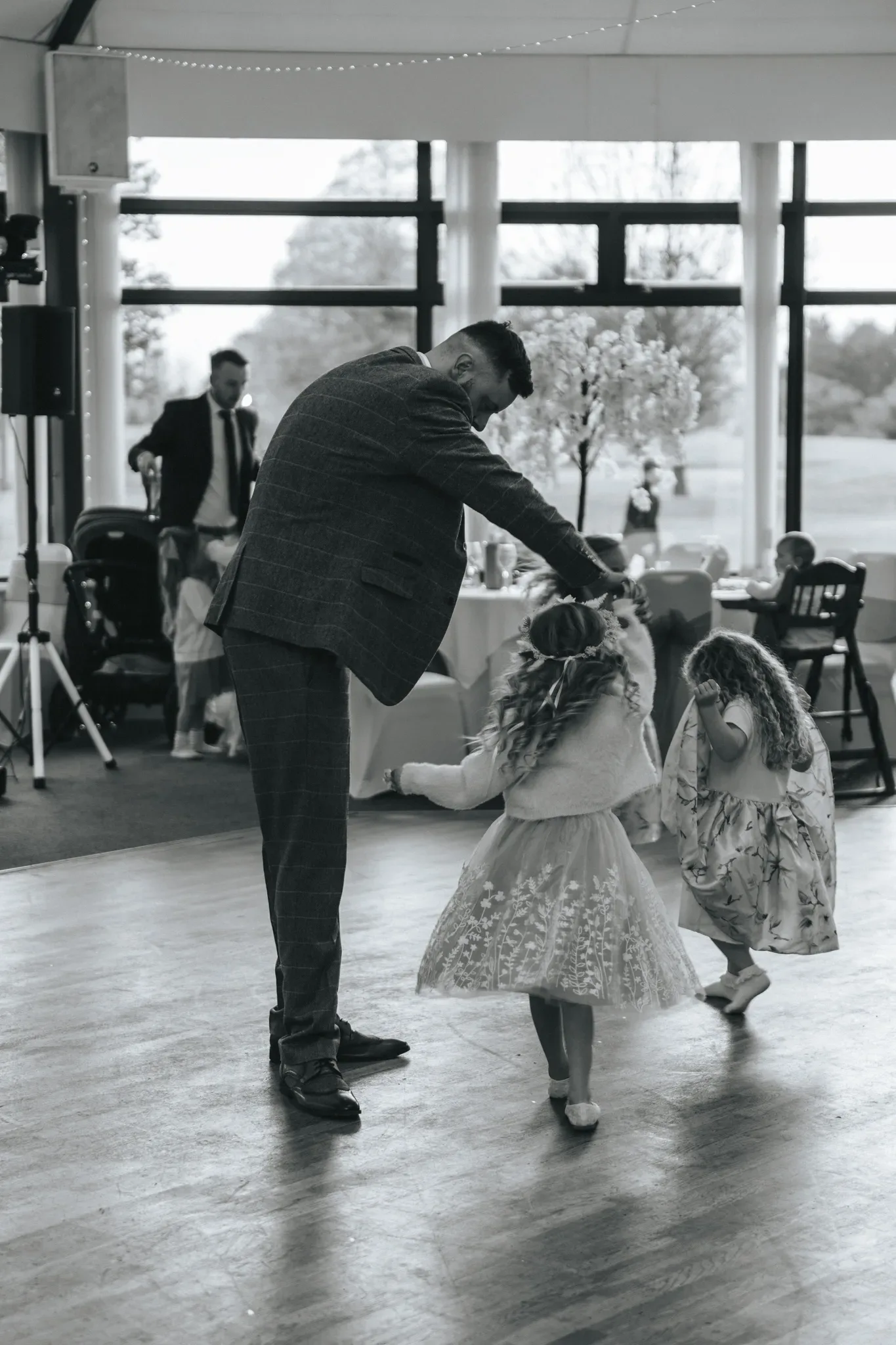 A man in a tailored suit playfully dances with two young girls in fancy dresses at an indoor event, likely a wedding. the setting features a bright room with large windows and a dance floor. the atmosphere is joyful and energetic.