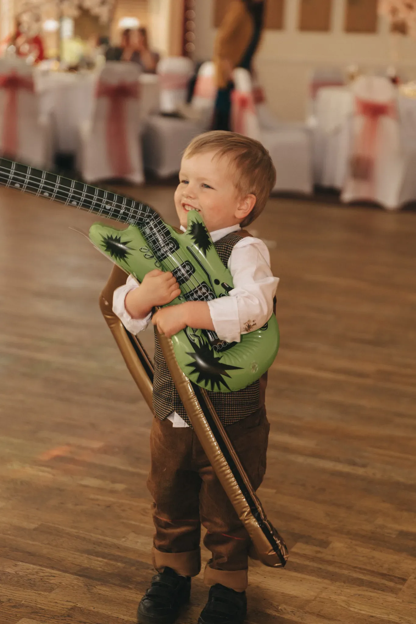 A young boy in a white shirt, vest, and brown pants, enthusiastically holding a large inflatable green guitar at a festive indoor event.