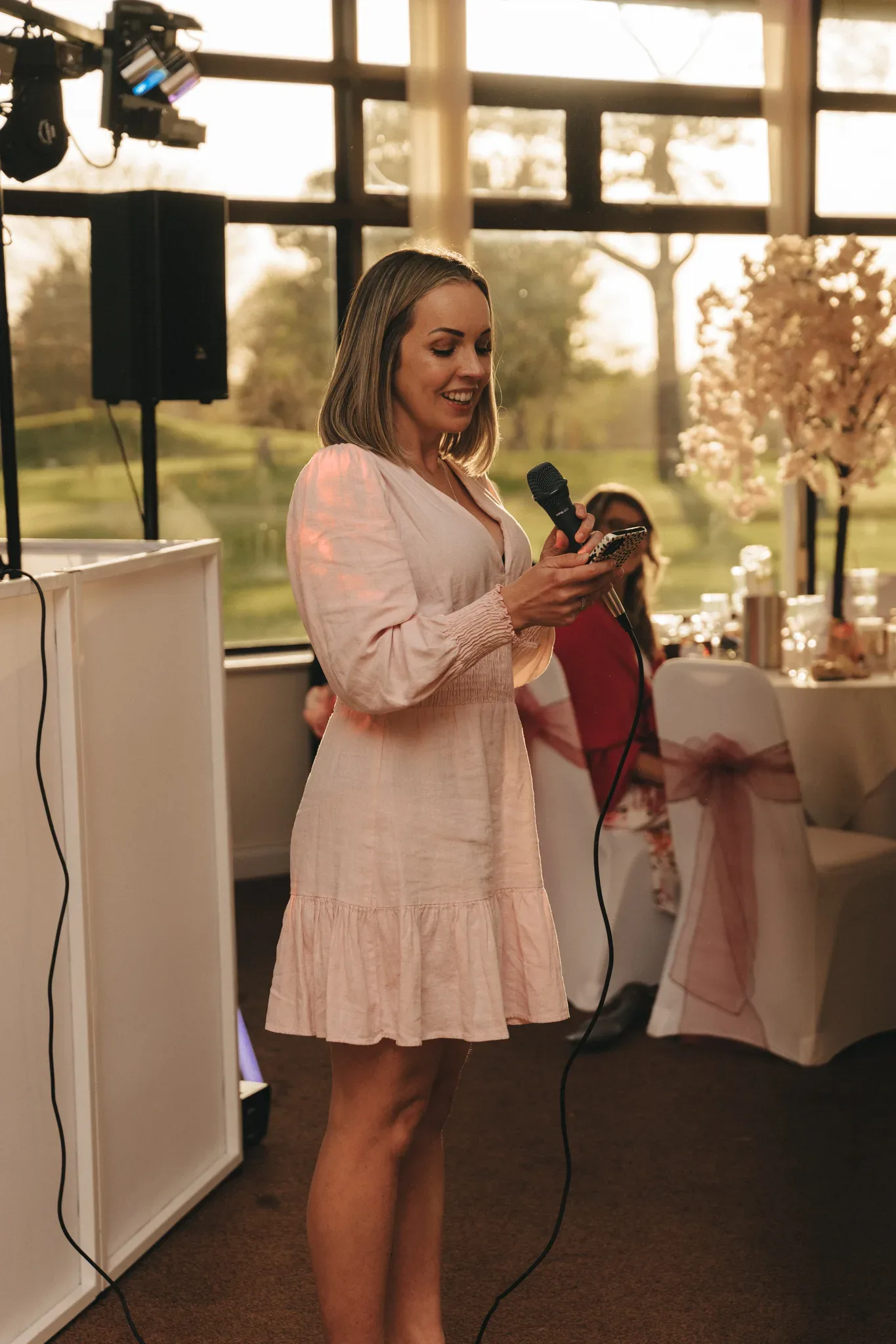 A woman in a light pink dress smiles as she gives a speech, holding a microphone and a smartphone. she stands indoors, with large windows and decorative seating in the background.