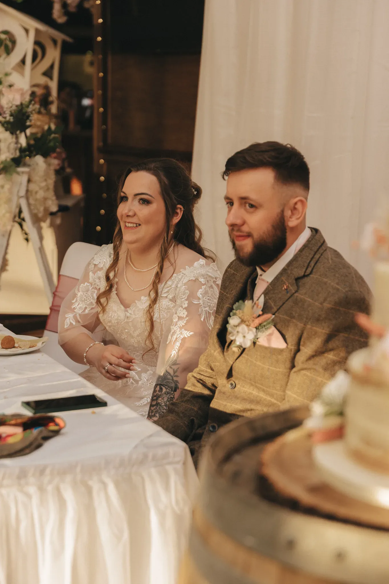 A bride and groom sit at a decorated wedding reception table, smiling while paying attention to a speech. the bride wears a white lace dress and the groom sports a tweed suit and flower boutonniere. a cake is visible in the foreground.