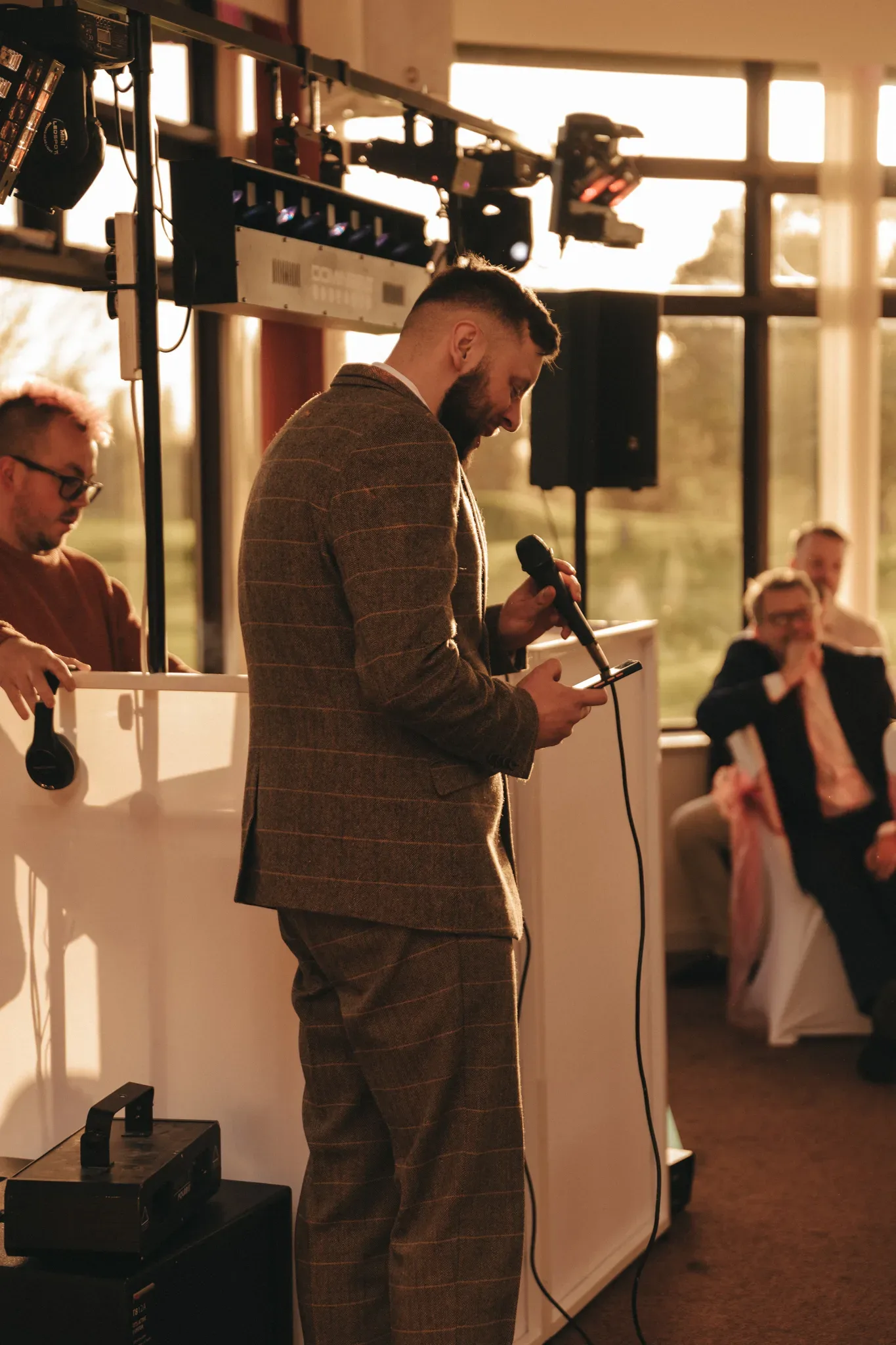 A man in a tweed suit delivering a speech at a podium with a microphone, with attentive guests in the background during a sunset event. bright sunlight casts warm glows through large windows.