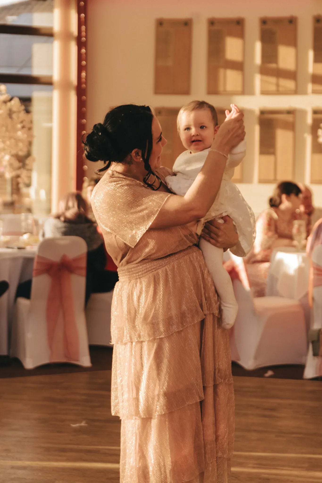 A woman in an elegant, shimmering pink dress holds a smiling baby in a white outfit at a festive event with decorated tables and chairs in the background.