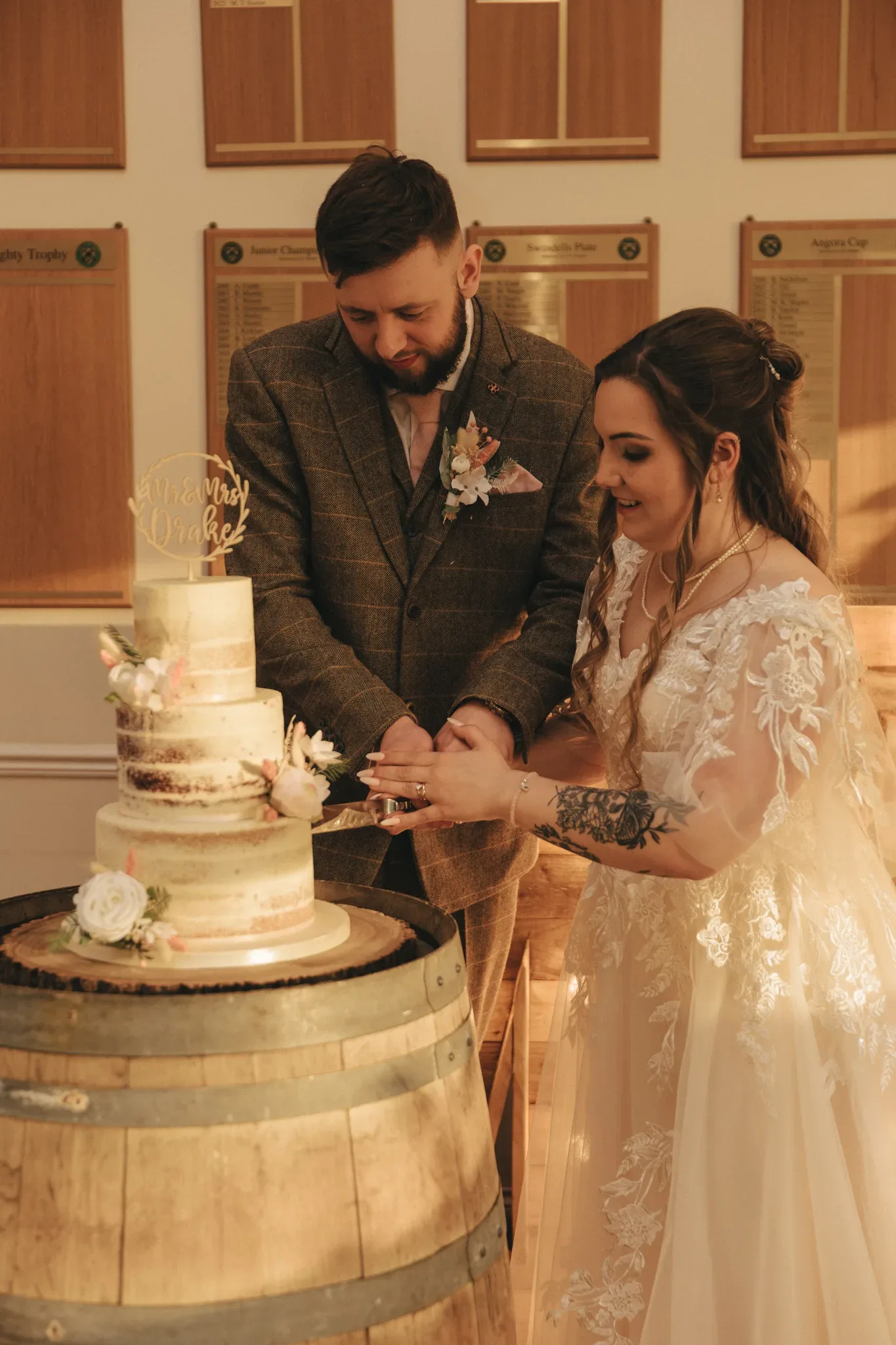 A bride and groom cut their wedding cake together. the bride, in a lace gown, and the groom, in a tweed suit, stand by a cake on a barrel in a warmly lit room. the cake adorned with flowers.