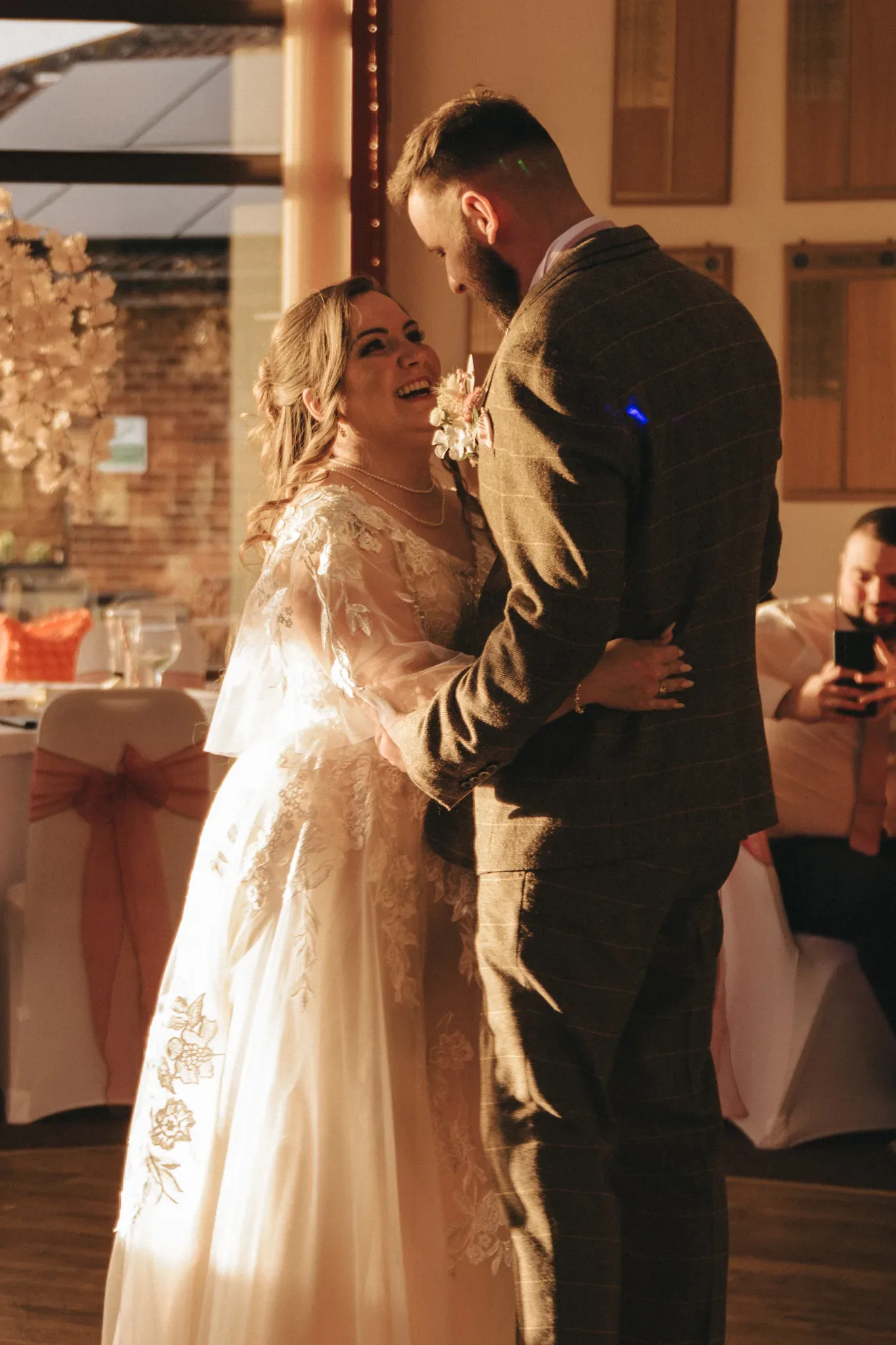 A bride and groom share a joyful dance under warm lighting at their wedding reception. the bride, in a lace-detailed dress with sheer sleeves, smiles up at the groom, who is dressed in a tailored tweed suit.