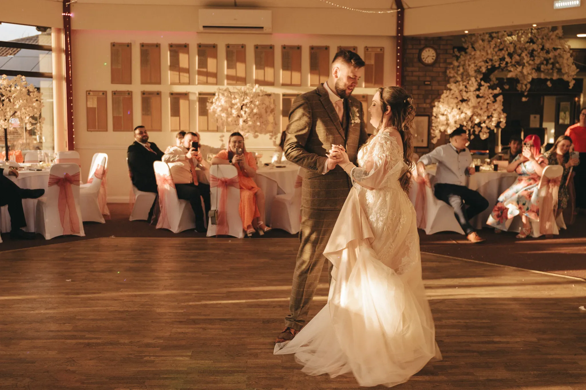 A bride and groom share a dance in a warmly lit hall adorned with white tablecloths and centerpieces of blossoming trees. guests seated around observe the intimate moment. the groom wears a checkered suit, and the bride a white gown.