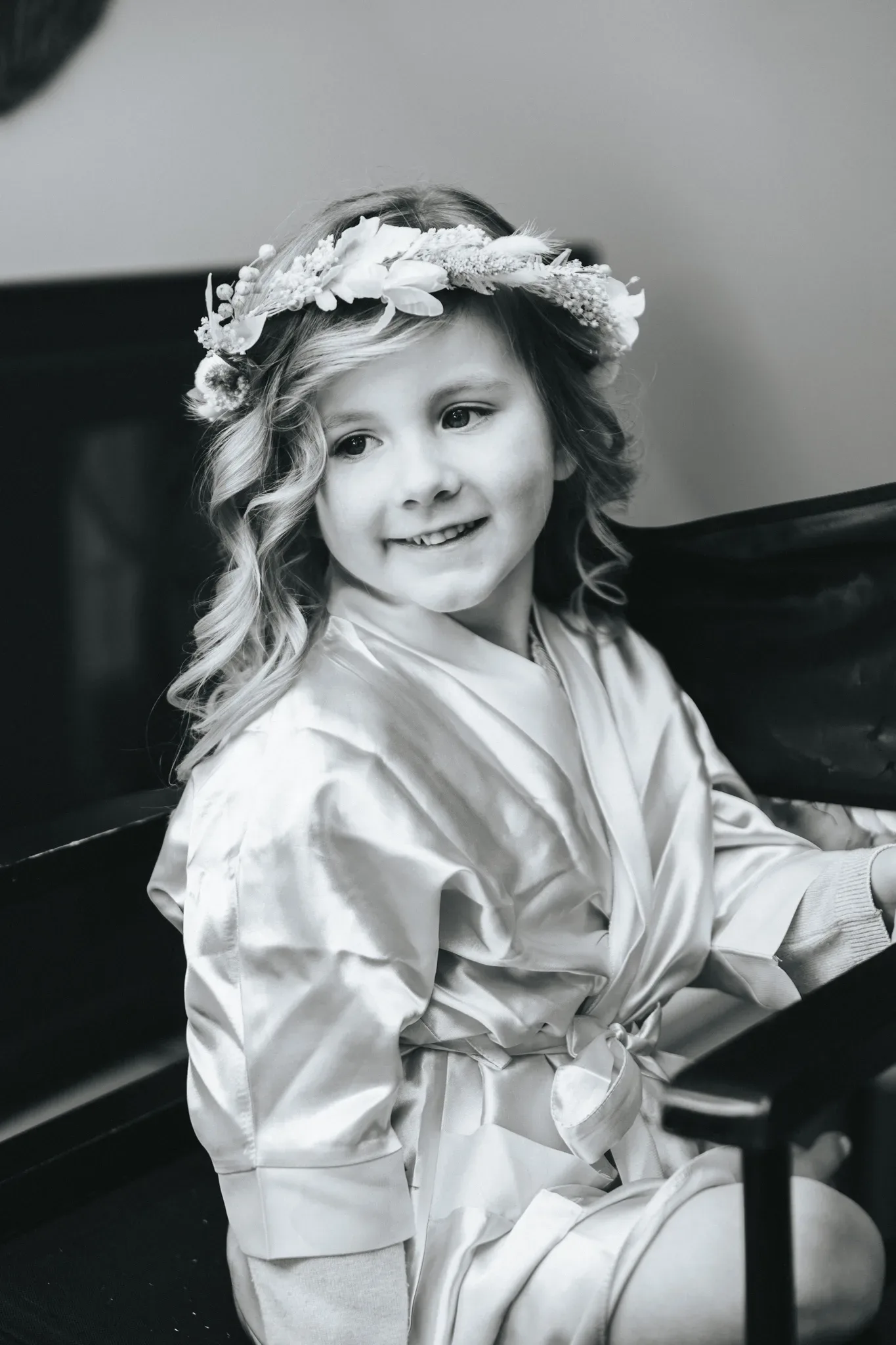 A black and white portrait of a young girl with curly hair, wearing a silk robe and a floral hat, sitting beside a piano and smiling gently at the camera.