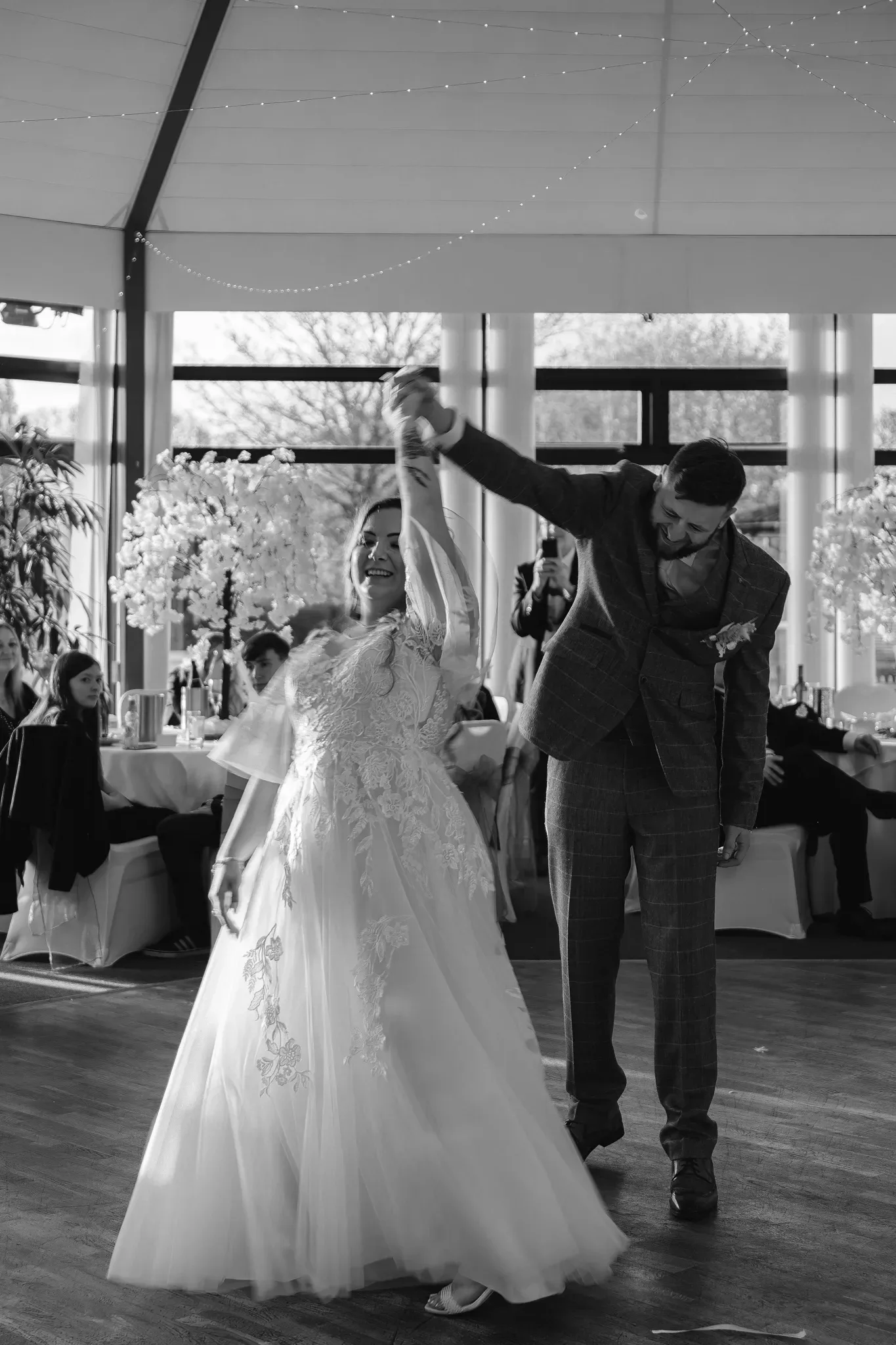 A joyful bride and groom share their first dance, the bride in a lace gown and the groom in a tweed suit. they dance under string lights in a room with guests seated around them, captured in black and white.