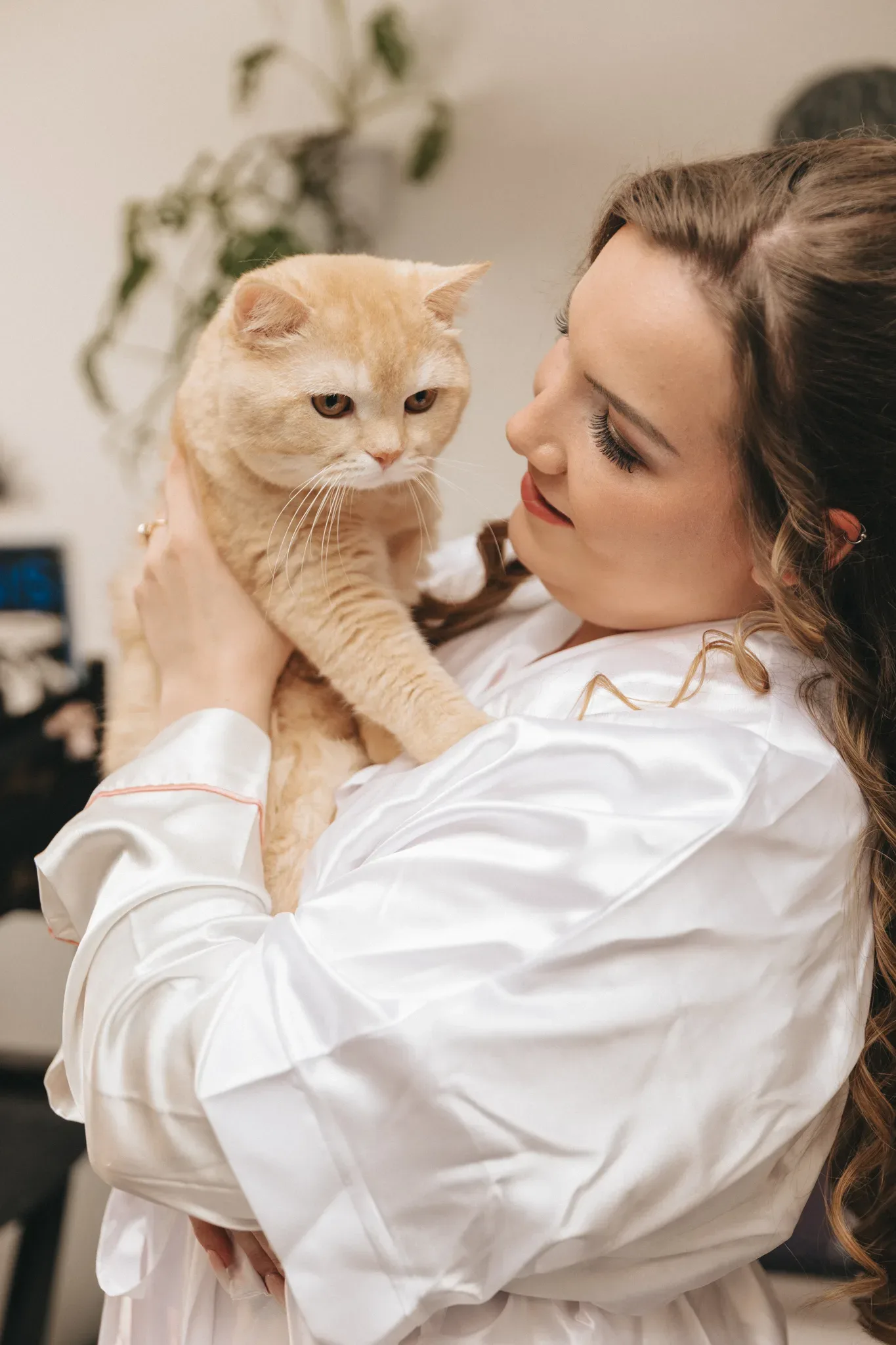 A woman in a white robe holds a chubby orange cat with a displeased expression. they are indoors, with soft focus on a cluttered background with plants.