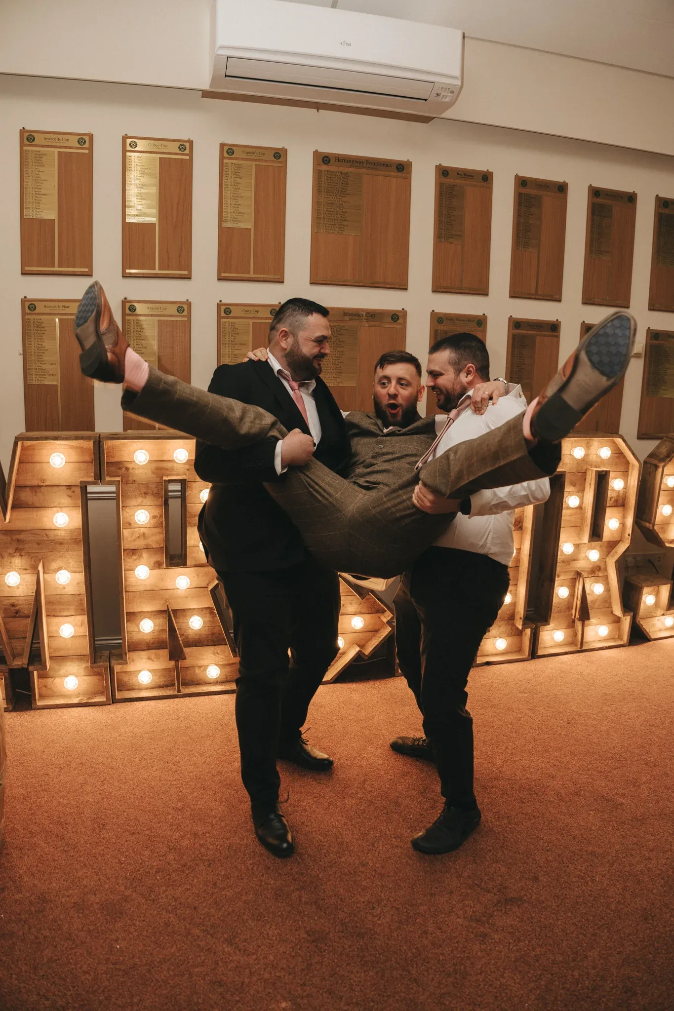Three men in a celebratory setting, playfully lifting a fourth man horizontally across their arms. they are all dressed in formal attire, with large lit "bar" letters in the background, adding a festive ambiance.