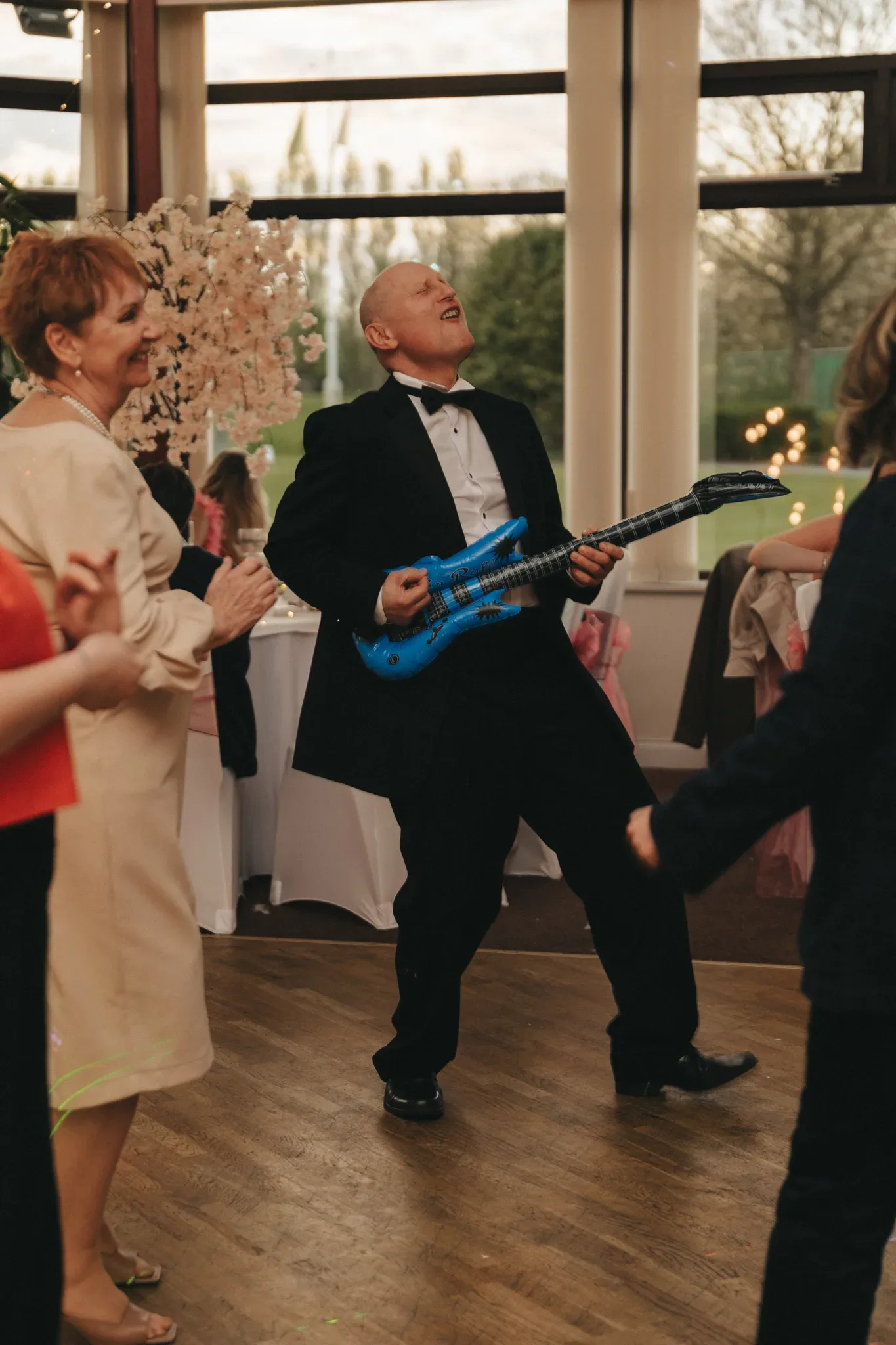 An elderly man joyfully plays a blue toy guitar at a party. he's dressed in a formal black suit and is surrounded by clapping guests, including an amused elderly woman, in a room with large windows and pink decor.
