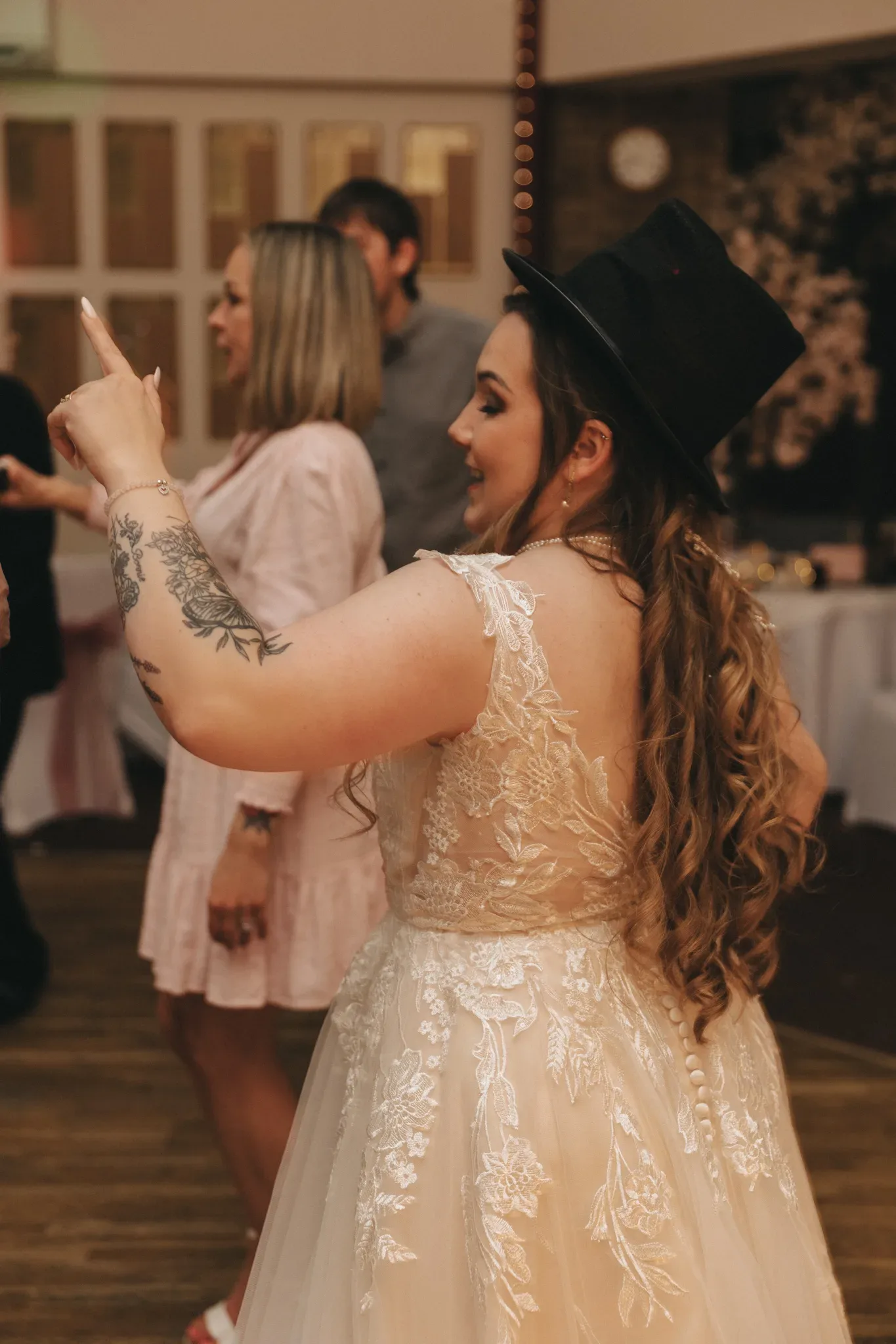 A woman in a detailed white lace dress and a black top hat interacts with her phone, likely taking a photo, at a lively indoor gathering with other guests chatting in the background.