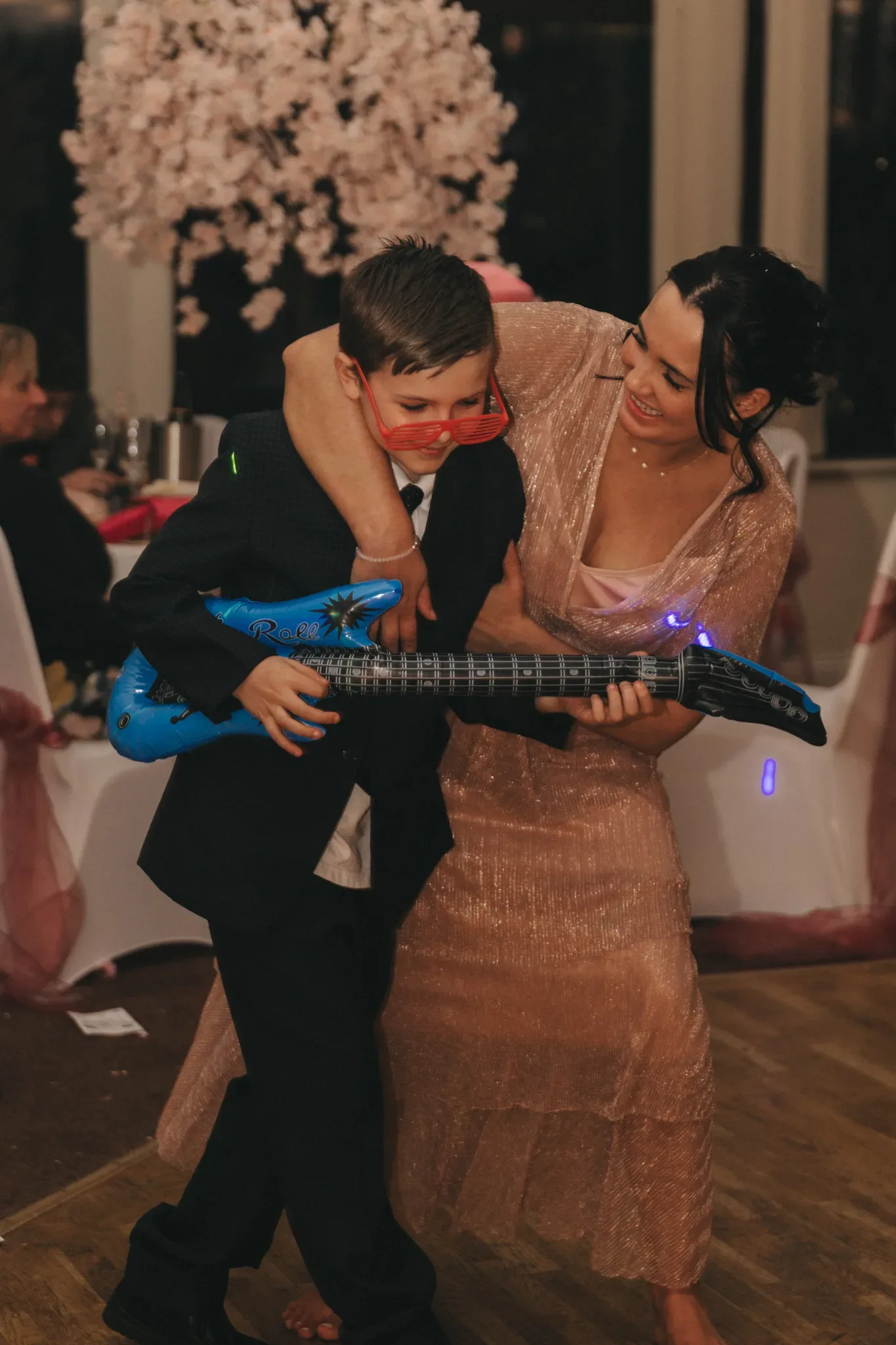 A woman and a young boy laugh together at a party, with the boy playfully pretending to play a blue toy guitar. the woman, dressed in a shimmering long dress, gently adjusts his suit jacket. elegant decor and seated guests in the background.