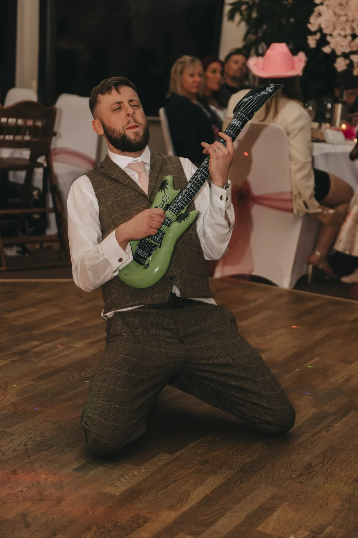 A man in a vest and tie kneels while playing a small green toy guitar, visibly enjoying himself at an indoor event with guests seated in the background.