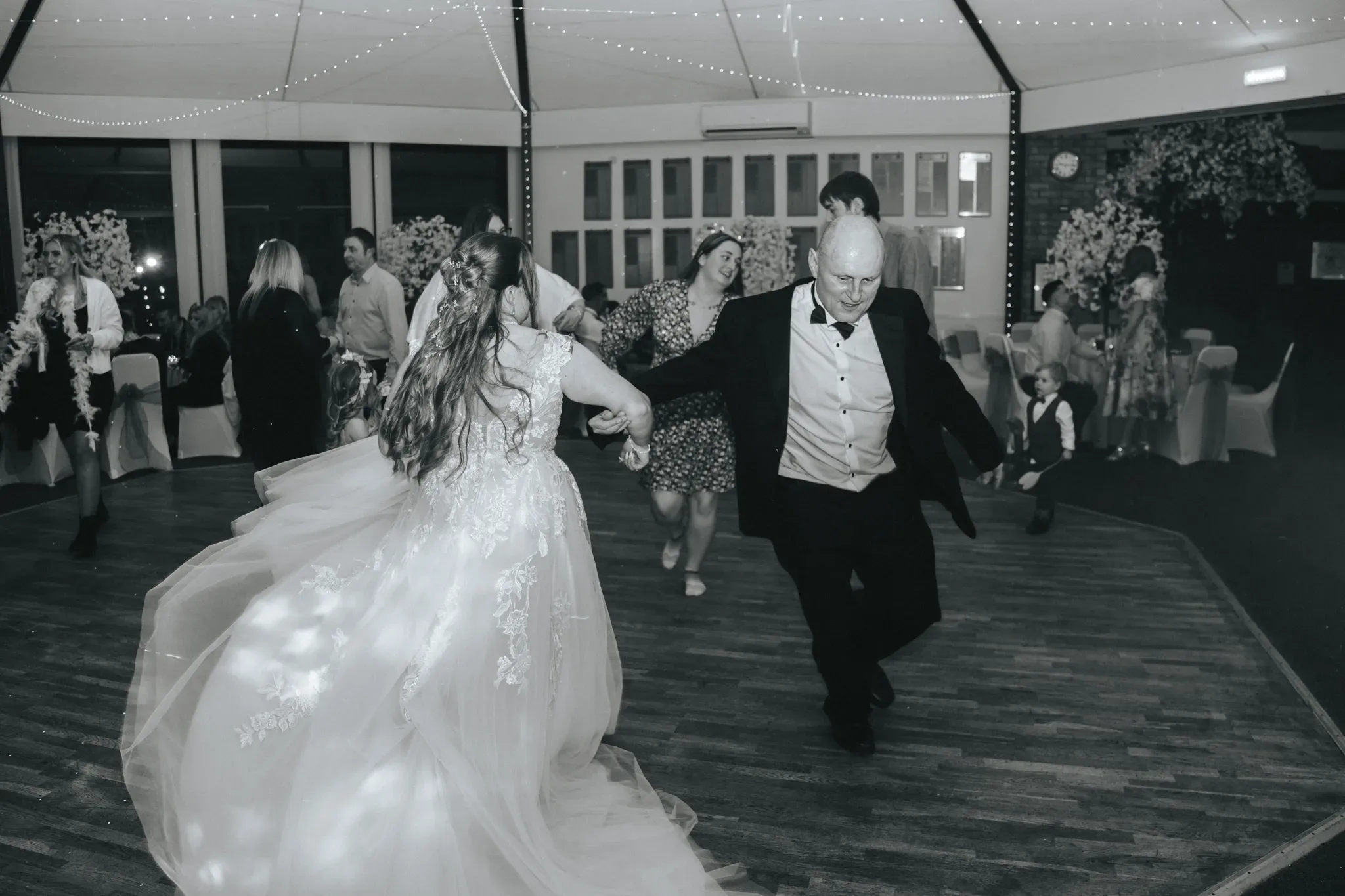 A bride and an older man dance joyfully at a wedding reception, surrounded by guests. the bride's detailed gown flows behind her as she moves, and string lights hang above the dance floor, enhancing the festive atmosphere.
