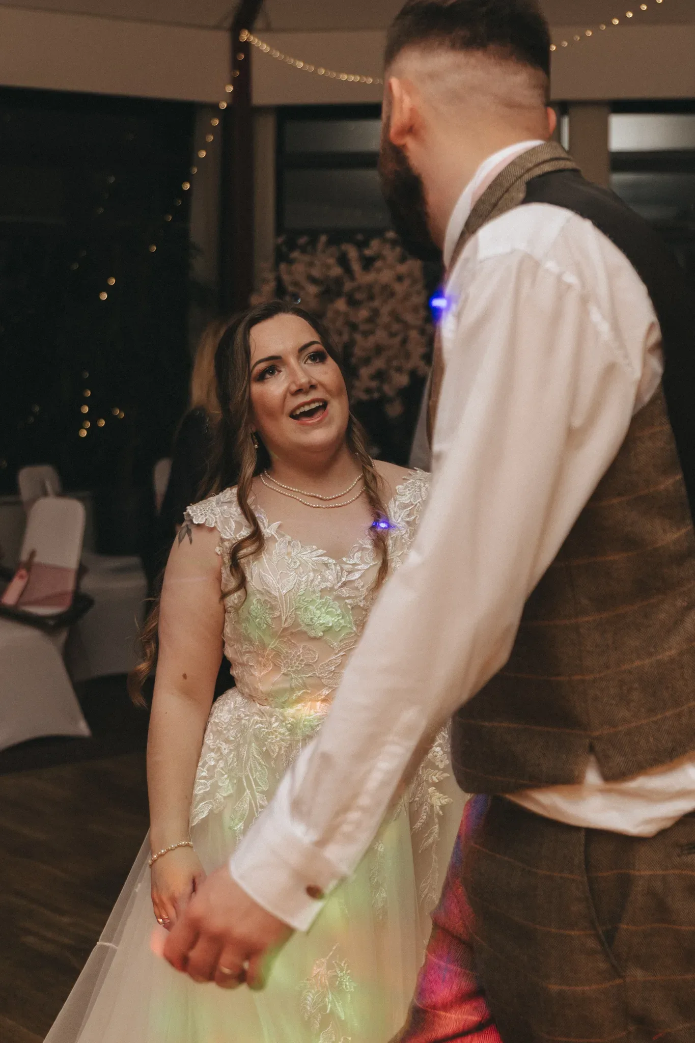 A bride with long wavy hair and a detailed dress smiles adoringly at a groom in a tweed vest. they hold hands while dancing, with festive lights and a warmly lit venue in the background.