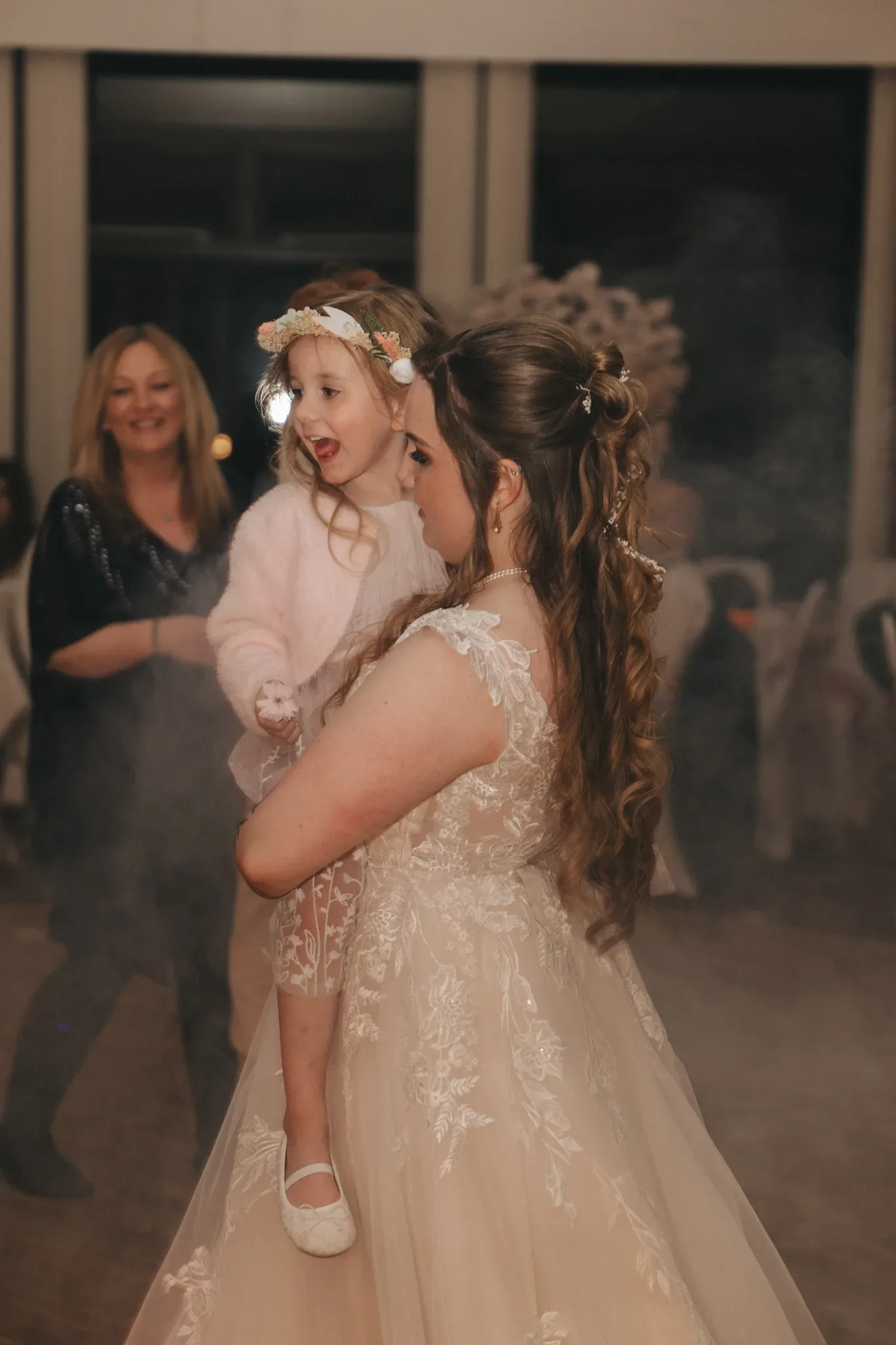 A woman in a lace wedding dress holds a young girl wearing a white dress and floral headband. they are surrounded by guests in a dimly lit room with visible joy and celebration.