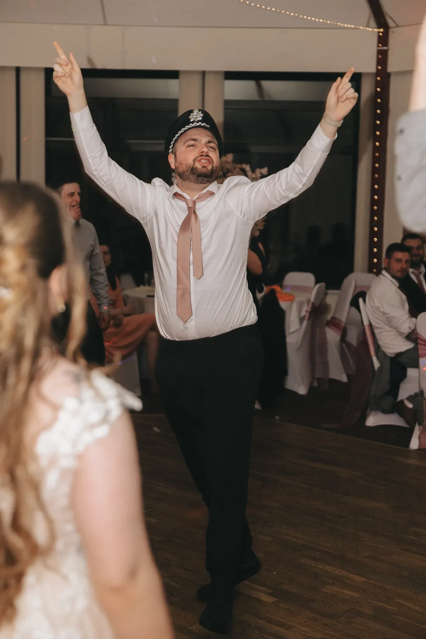 A man at a wedding reception, dressed in a formal shirt and tie, joyfully dances with his arms raised. guests seated around him are watching and smiling, creating a festive atmosphere.