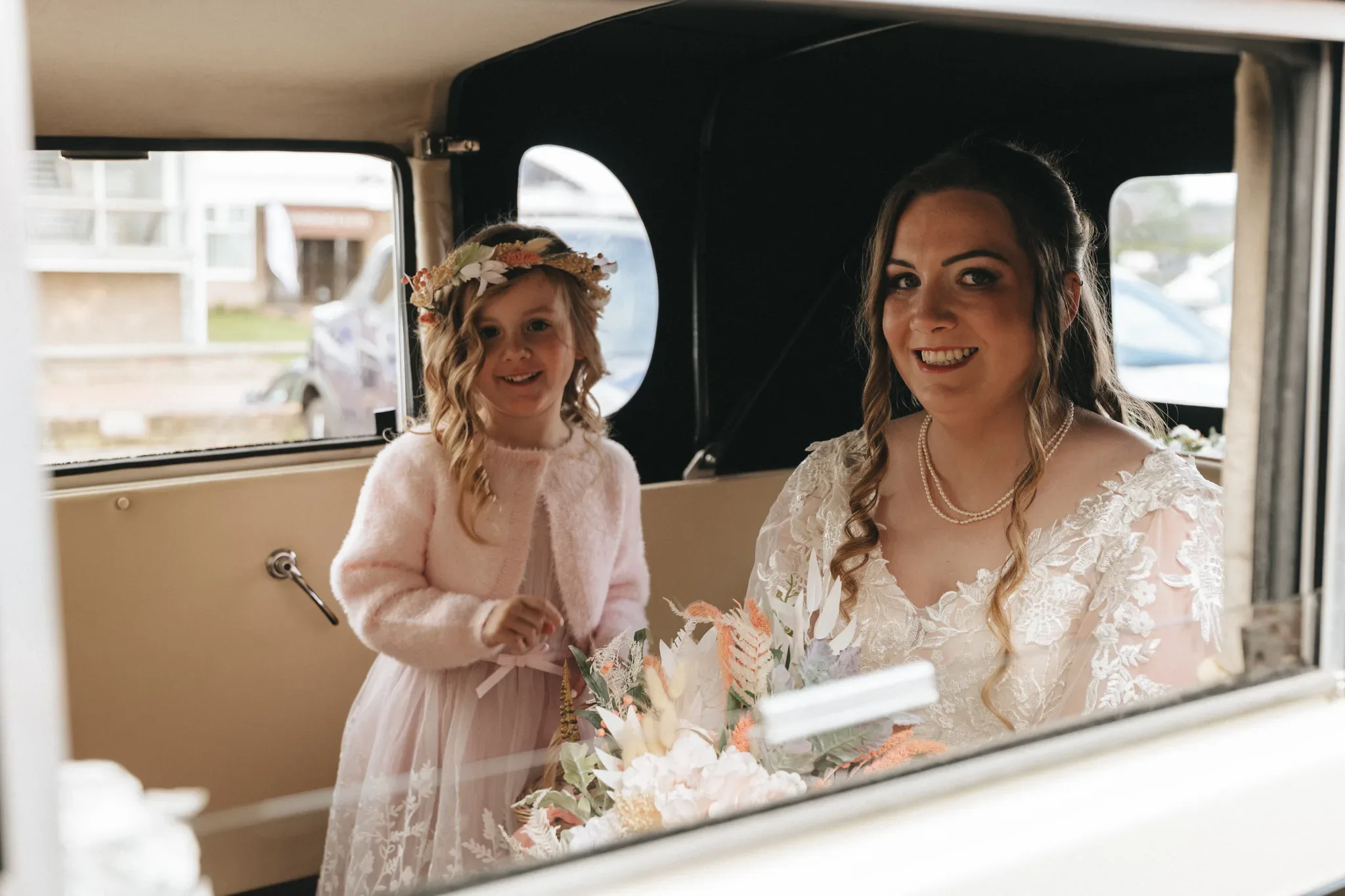 A bride and a young girl in a flower crown smile inside a vintage car, holding a bouquet. the bride wears a lace dress and the child a pink coat. they appear joyful, framed by the car window.