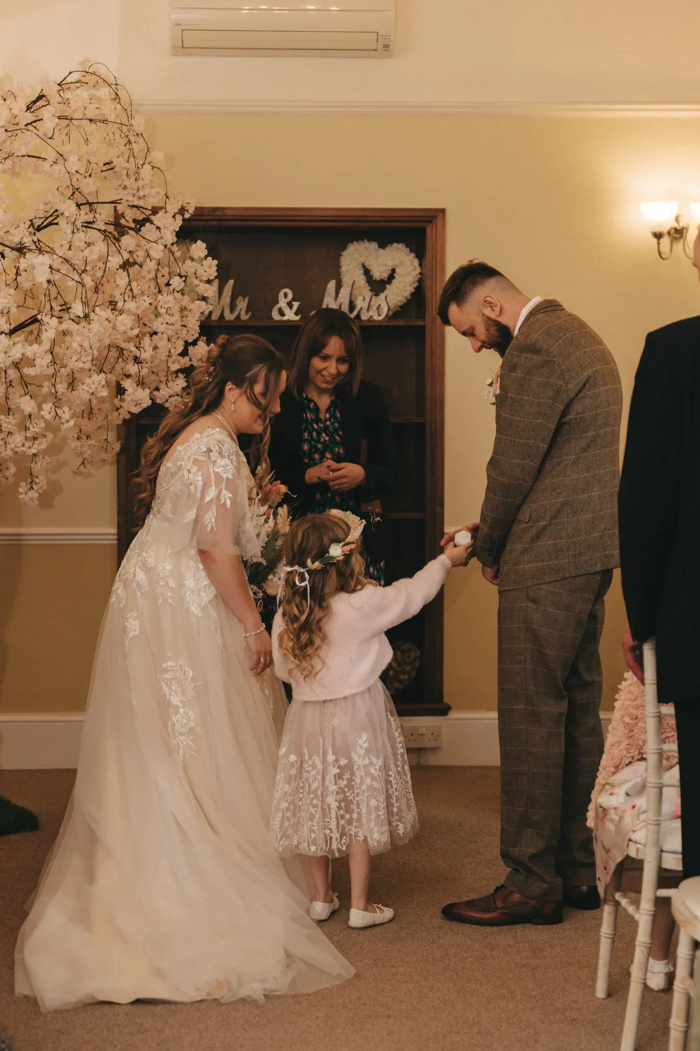A bride in an embroidered gown hands a flower to a young girl, while a groom and another woman watch. they are in a warmly lit room decorated for a wedding, with a "mr. & mrs." sign on the door.