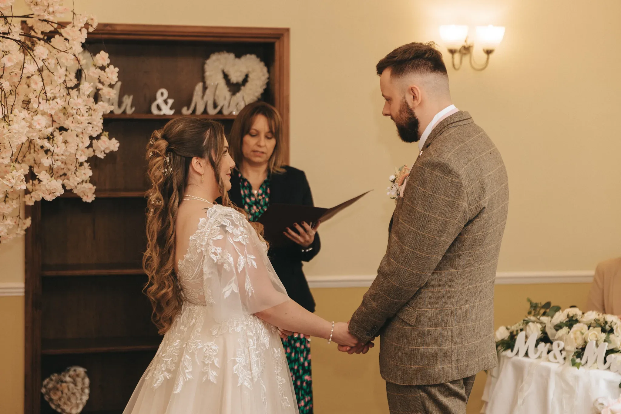 A bride and groom hold hands during a wedding ceremony, officiated by a woman in the background. the bride wears a white dress with lace sleeves, and the groom is in a tweed suit. a floral arch and "mr & mrs" sign decorate the room.