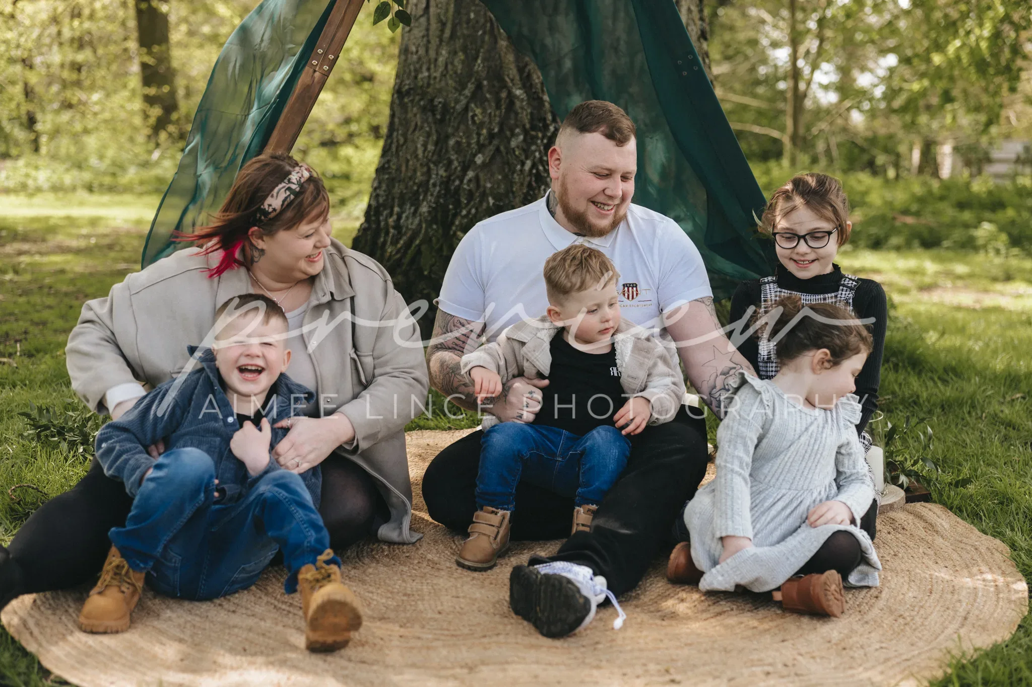 A joyful family of six poses for a Gabrielle photography session on a circular mat under a tree with a tent, laughing together. The parents and four children, including a toddler and an infant, enjoy